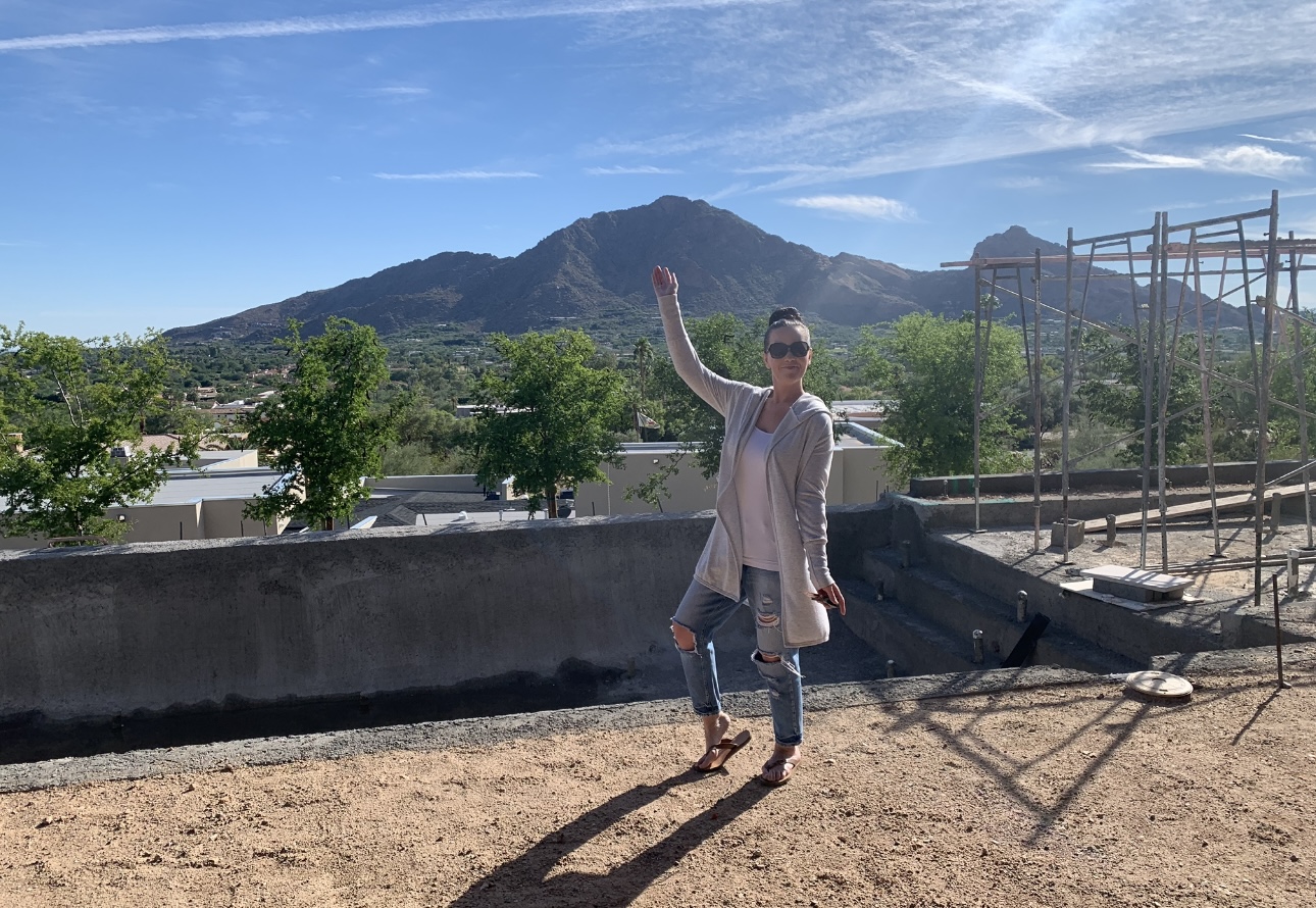 Person standing outdoors with mountains and trees in background, raising one arm, wearing sunglasses and a light coat.