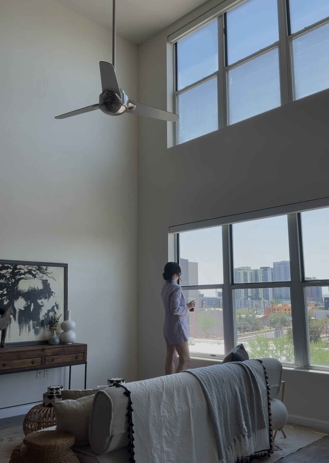 Person standing by large windows in a high-ceiling room with a ceiling fan and a sofa, holding a drink.