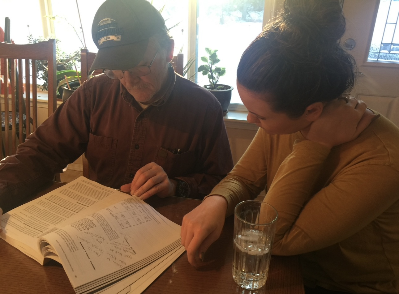 Two people sit at a table reading a large open book, with a glass of water nearby, in a well-lit room.