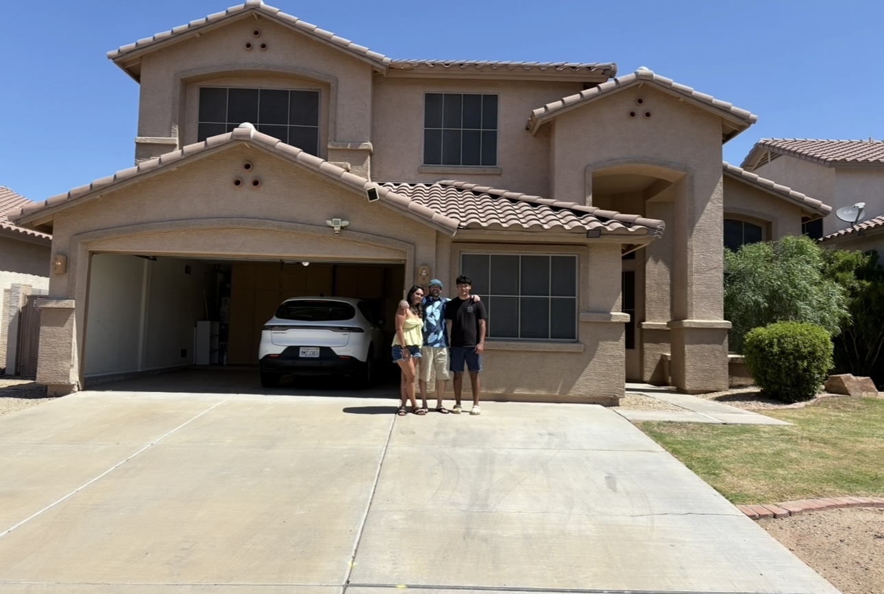 Three people stand in front of a two-story house with a garage and a white car inside, on a sunny day.