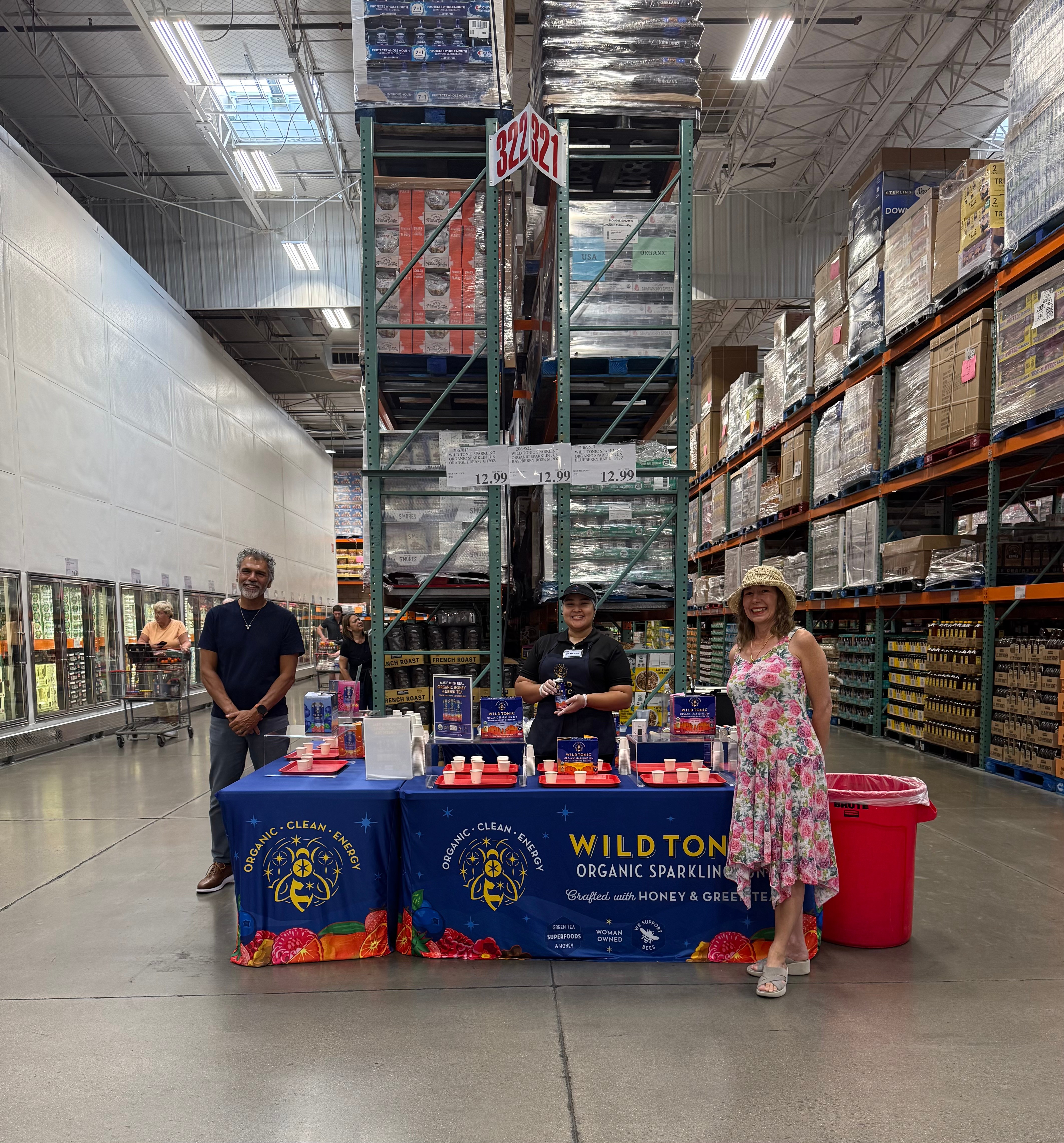 Three people standing behind a table with products in a warehouse, with shelves and a tall storage rack behind them.