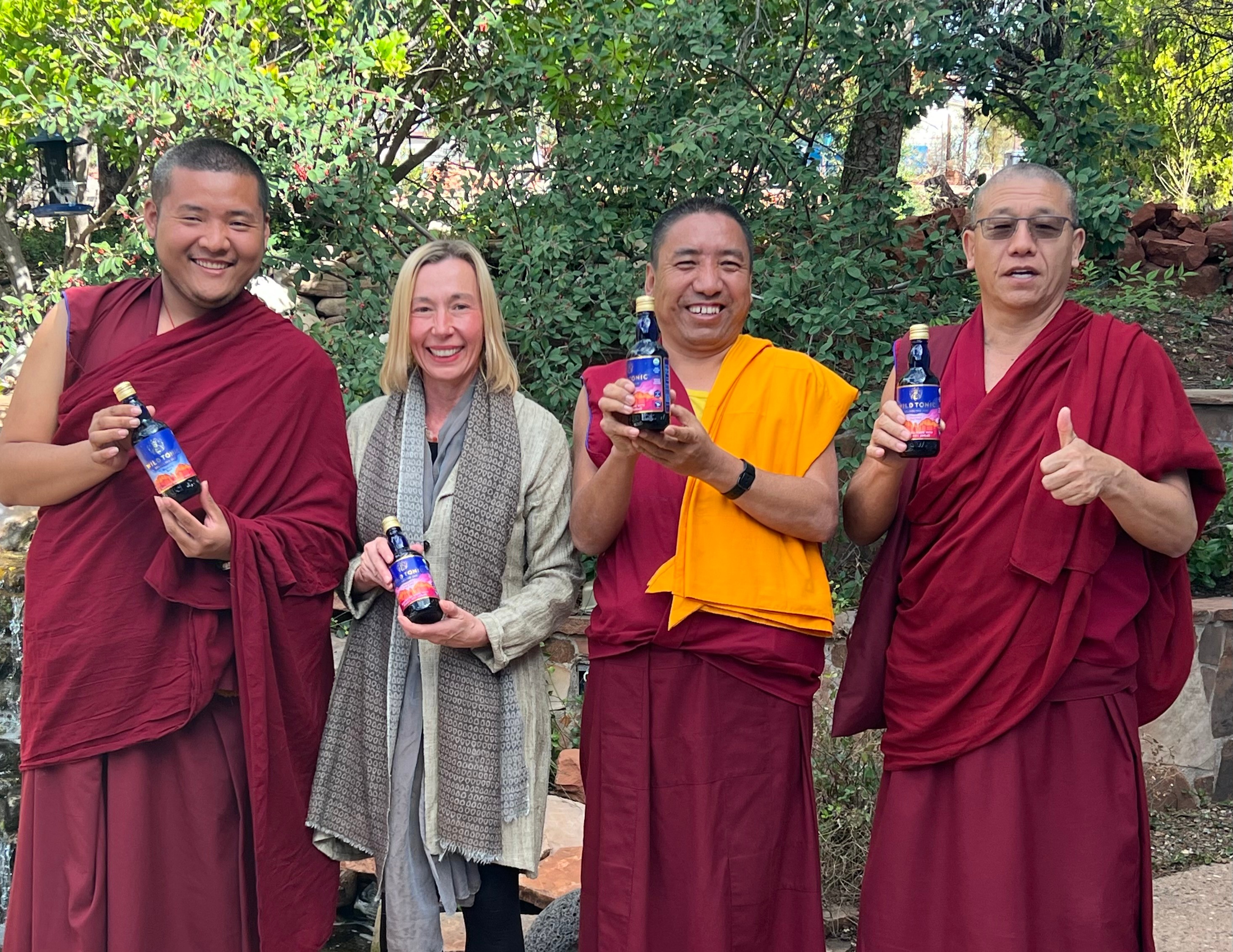 Four people, three monks in maroon robes and one woman in a gray coat, holding bottles outdoors with trees in background.