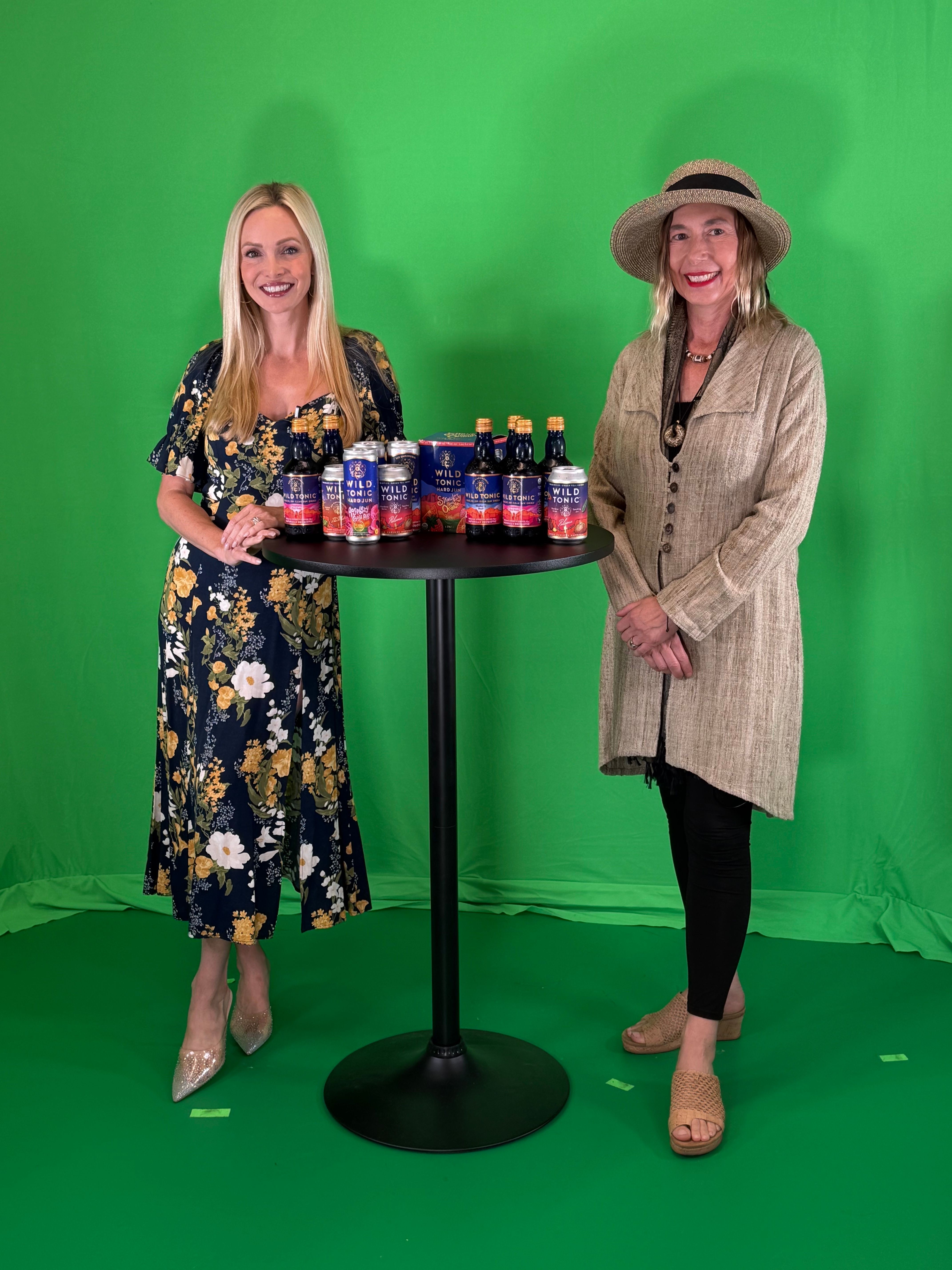 Two women stand in front of a green screen, one holding a tray of bottled drinks, both smiling, dressed in fashionable clothing.