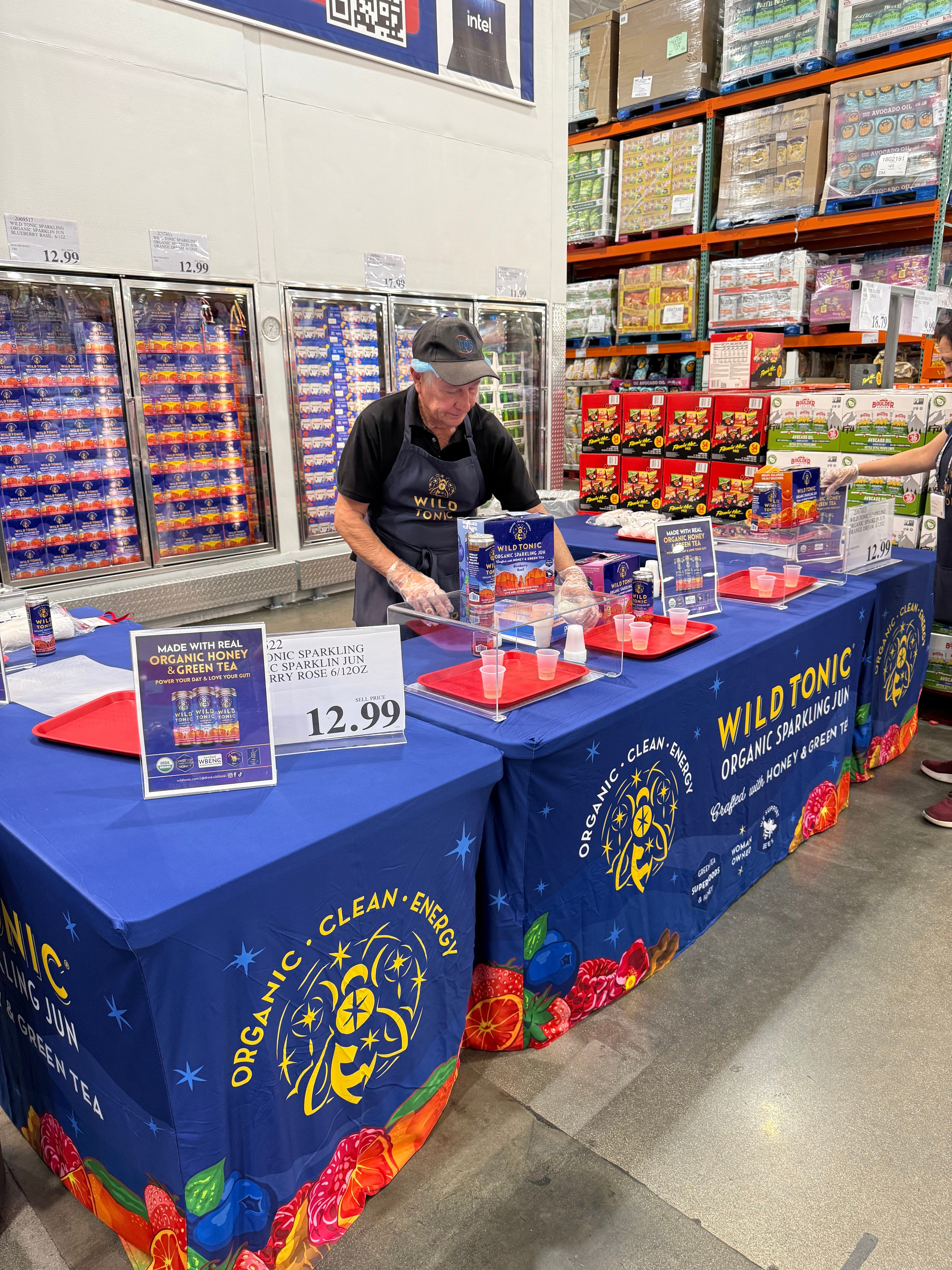 Person standing behind a table with books and promotional materials, shelves with books in the background, table covered with a blue cloth.