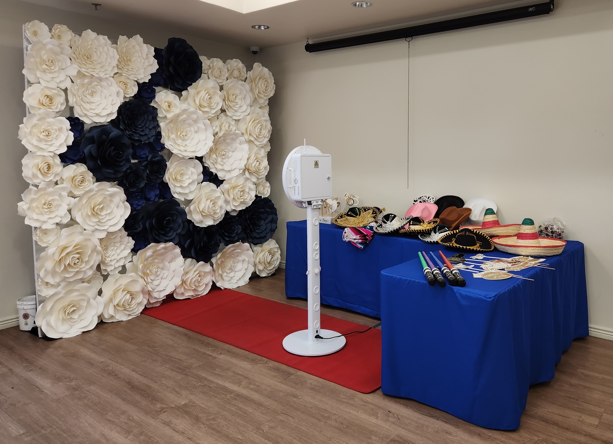 Display of hats and accessories on a blue table, with a large floral backdrop of white and dark flowers, in a room.