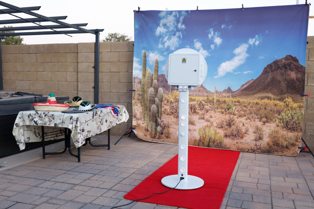 Photo of a photo booth setup outdoors with a desert landscape backdrop, a red carpet, and a white photo booth stand.