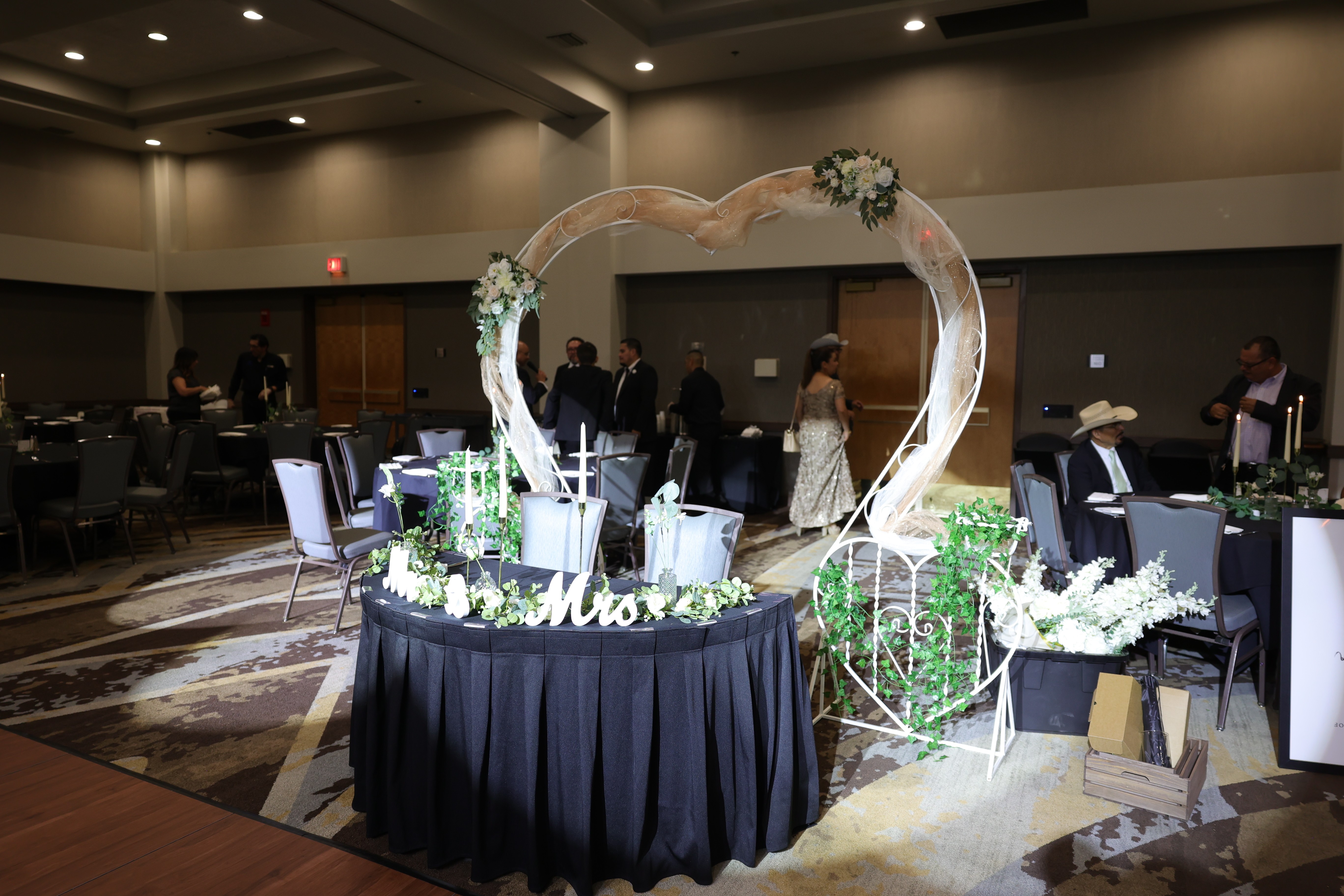 Round table with floral decorations and a heart-shaped arch in a banquet hall, with people in the background.