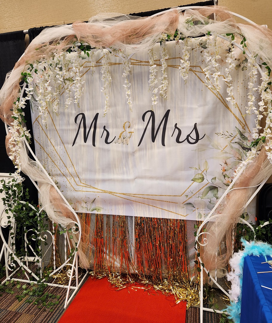 Wedding arch with floral decorations and a sign reading 'Mr & Mrs' on a red carpet, with a blue table nearby.