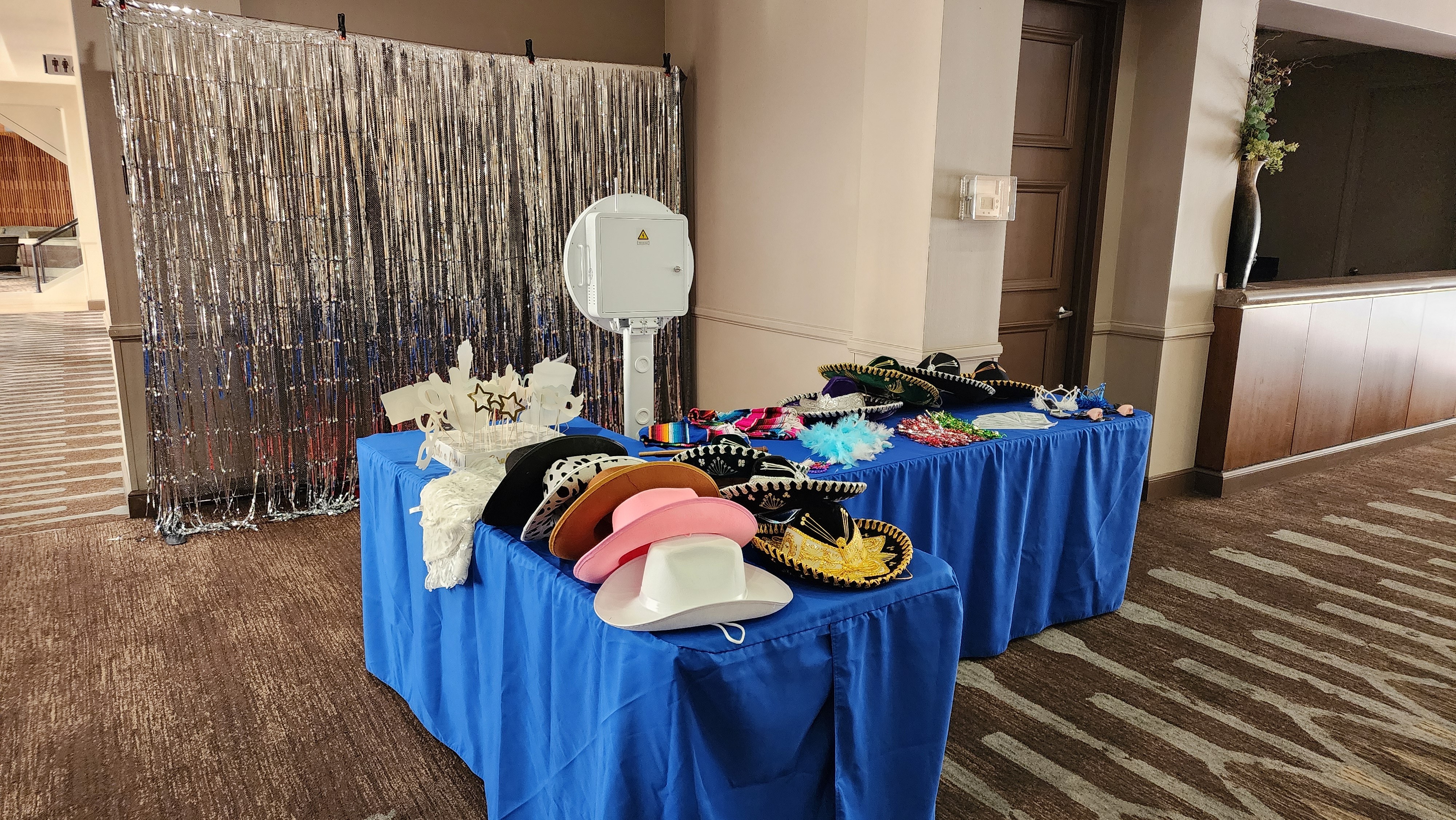 Table with hats and accessories covered with a blue cloth, set up indoors near a wall and decorative curtain.