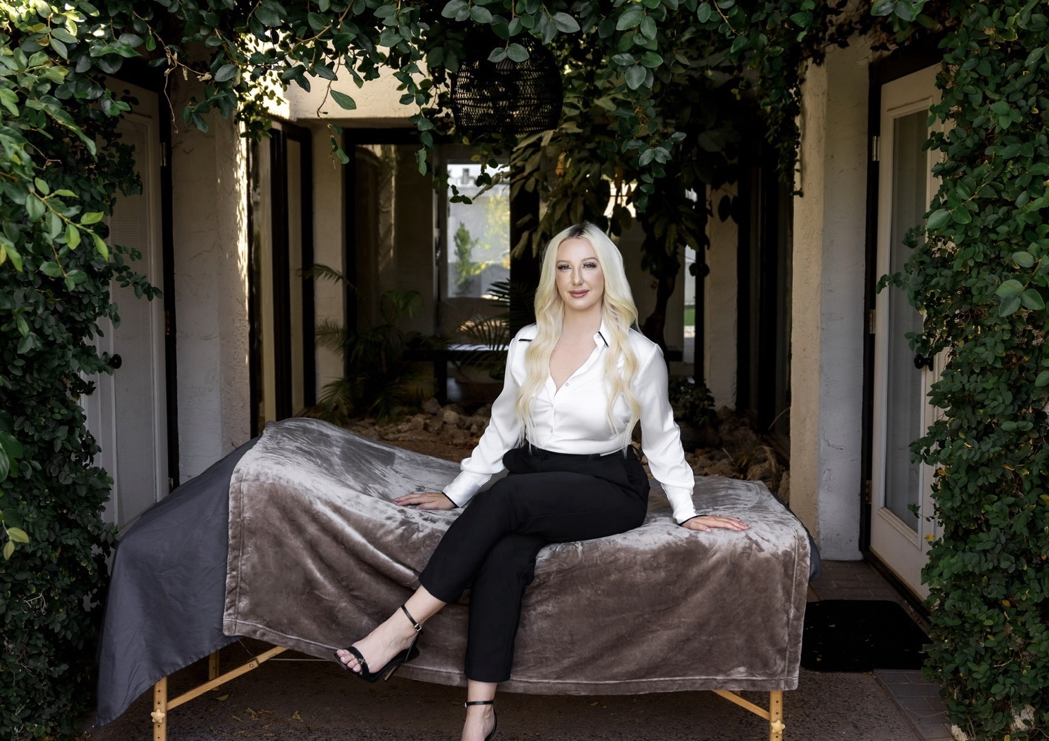 Woman with long blonde hair sitting on a velvet-covered bench outdoors, surrounded by greenery and trees.