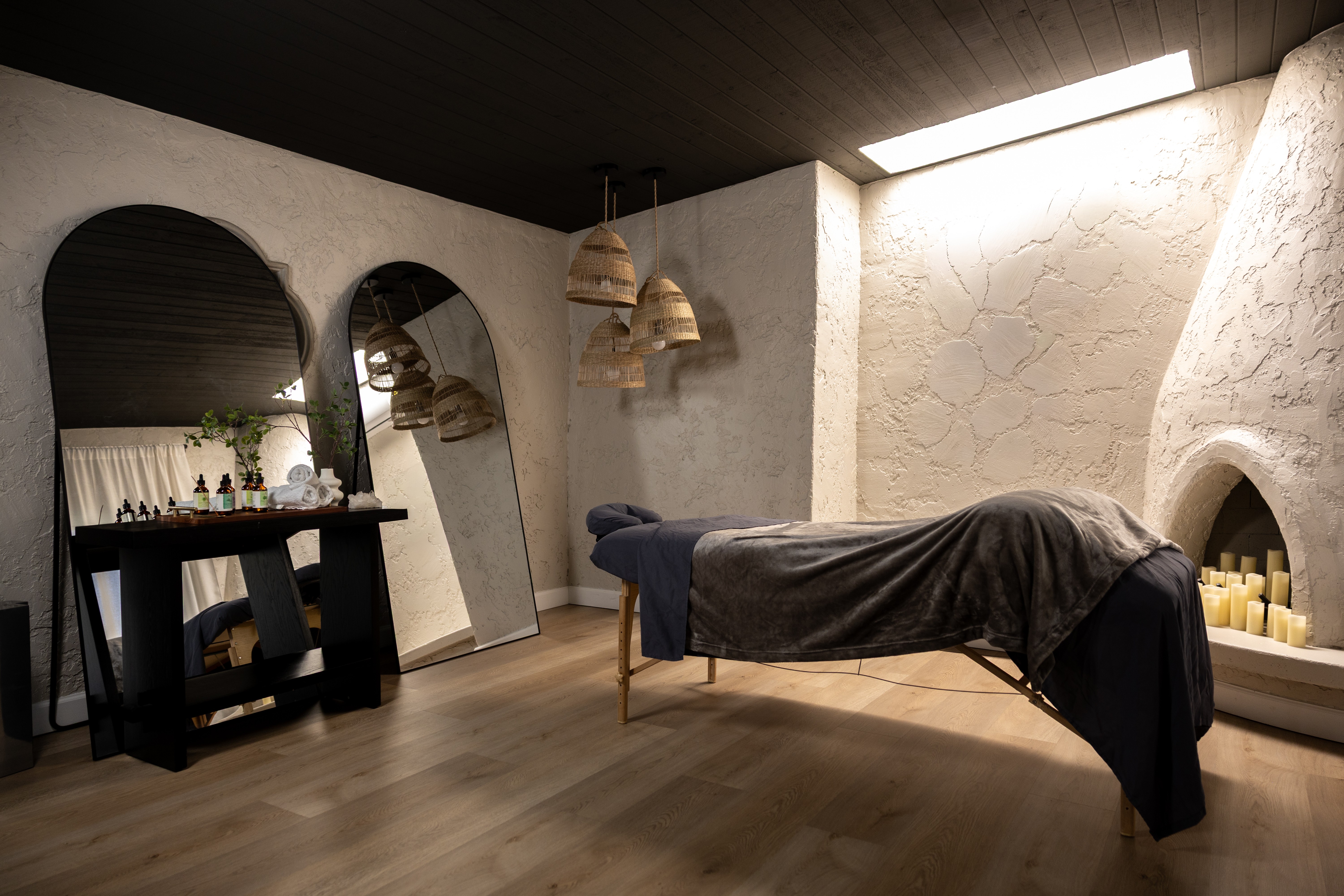 Massage table covered with dark cloth in a room with textured white walls, wooden floor, and hanging pendant lights.