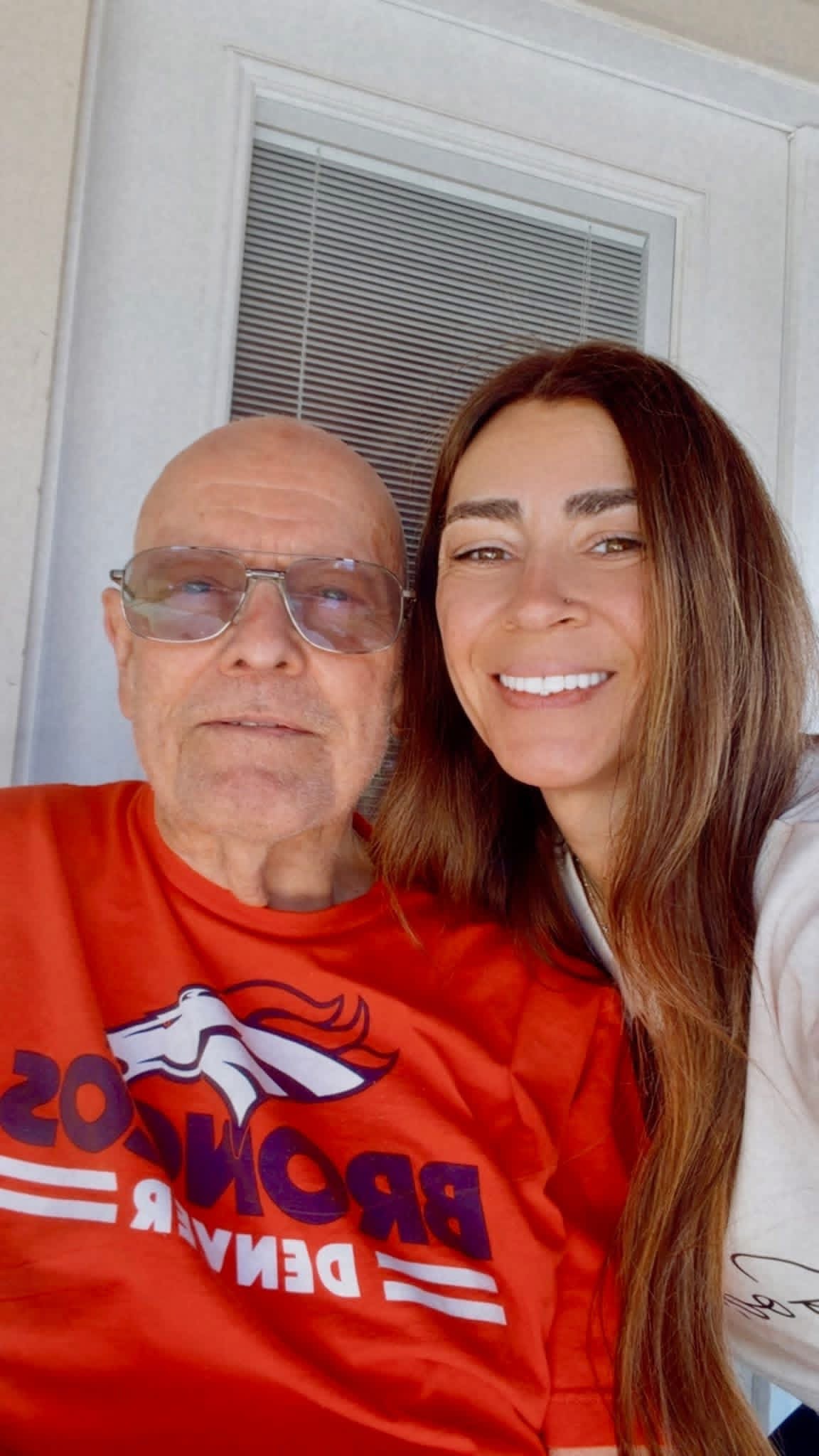 A woman with long brown hair and a man with glasses and a red Denver Broncos shirt, smiling together indoors.