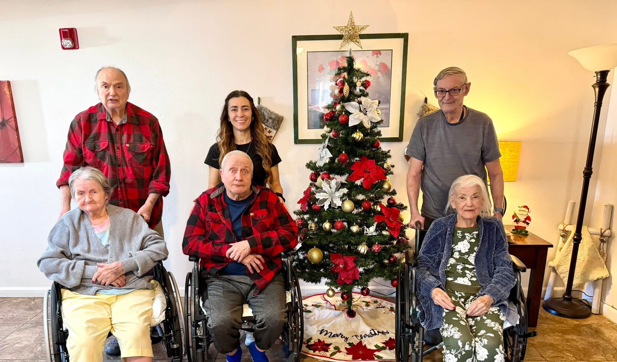 Six people, including elderly individuals in wheelchairs, gather around a decorated Christmas tree in a living room.