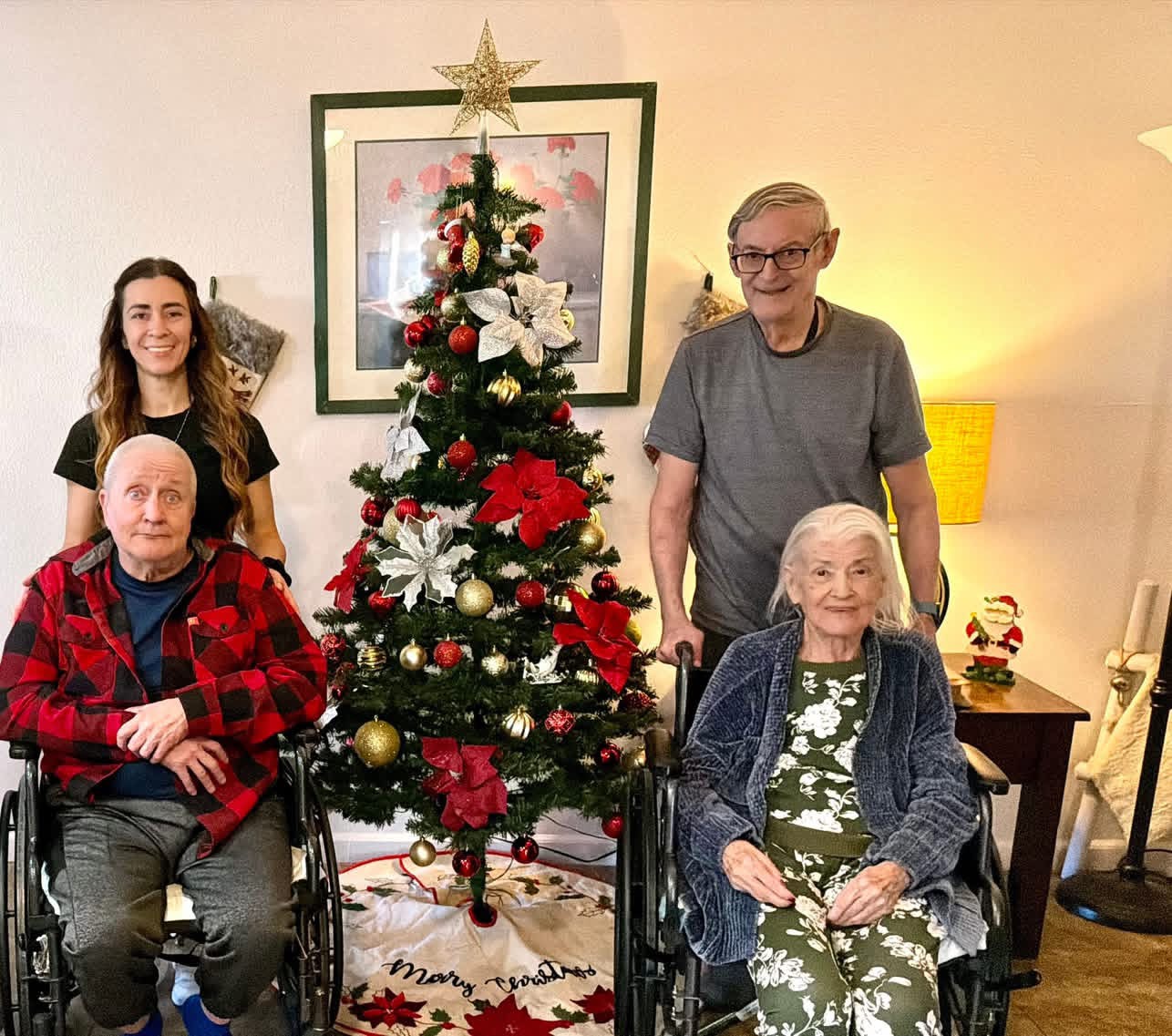 Four people, two in wheelchairs, stand around a decorated Christmas tree in a living room, smiling. A woman and a man stand behind, an elderly woman and a man in front. Christmas decorations and a framed picture are visible.