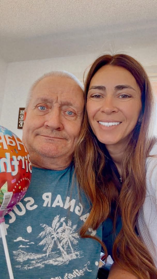 Two people smiling for a selfie indoors, one with long brown hair and the other with short gray hair, holding a colorful balloon.