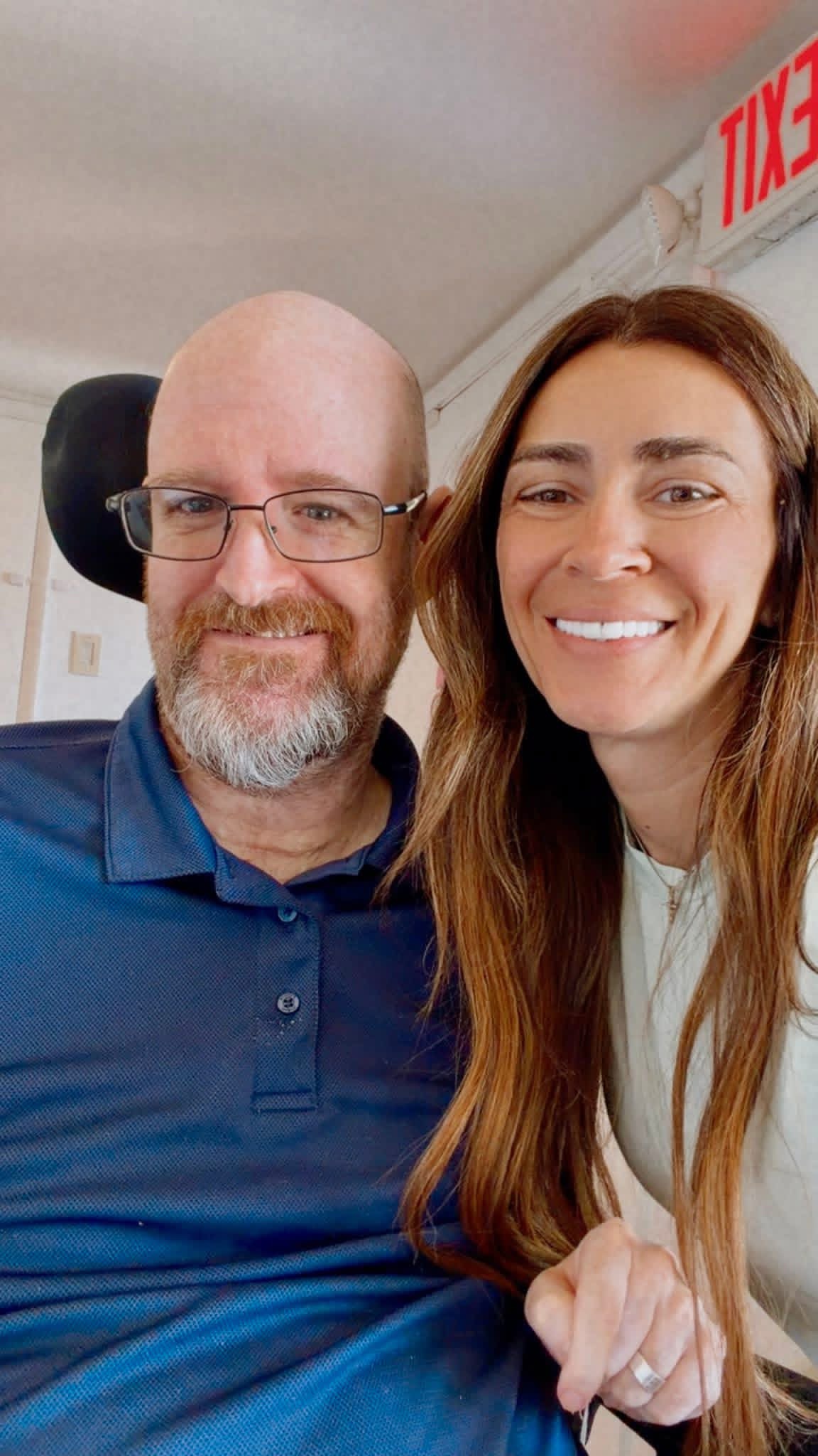 Two people smiling, one with glasses and a beard, the other with long hair, in an indoor setting with a red sign in the background.
