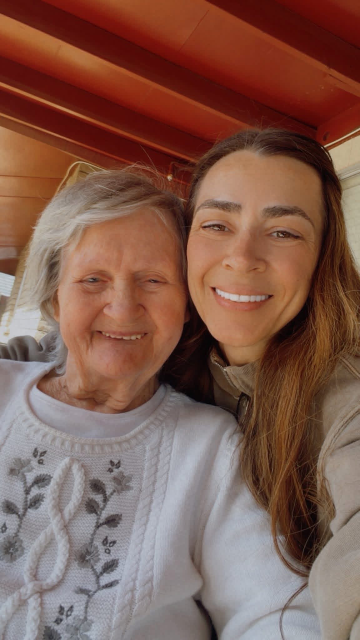 Two women smiling closely, one elderly with gray hair and the other younger with long brown hair, indoors with a wooden ceiling.