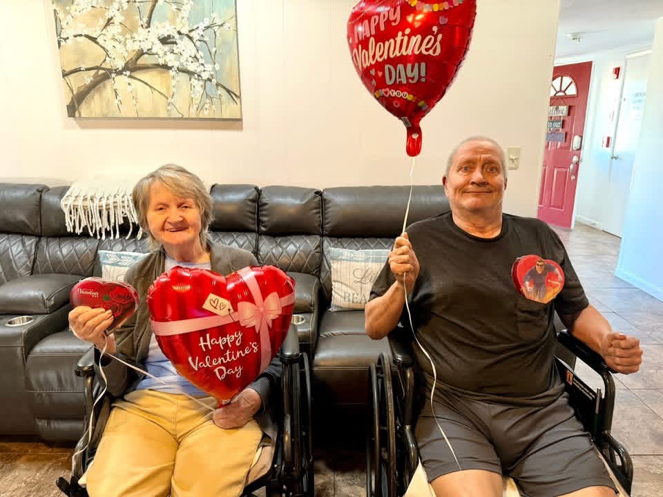 An elderly woman and a man in wheelchairs hold heart-shaped balloons and a large heart-shaped sign with Valentine's Day messages, sitting on a black leather couch in a living room.