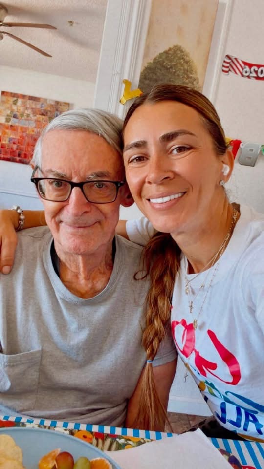Two women smiling, one older with glasses and gray hair, the other younger with long brown hair, in a bright room.
