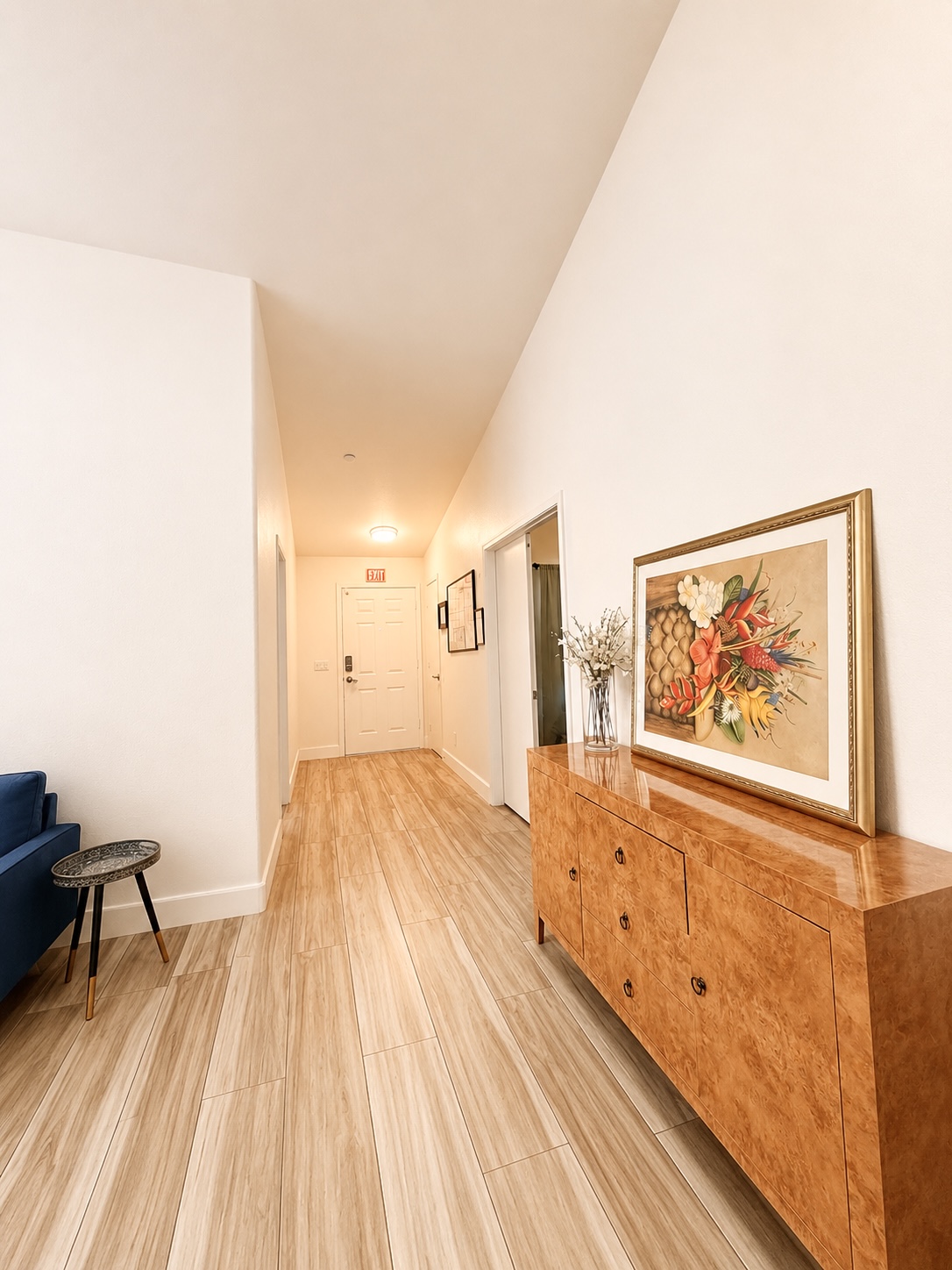 Long hallway with wooden floor, white walls, and a wooden sideboard with framed artwork and a vase, leading to a door.