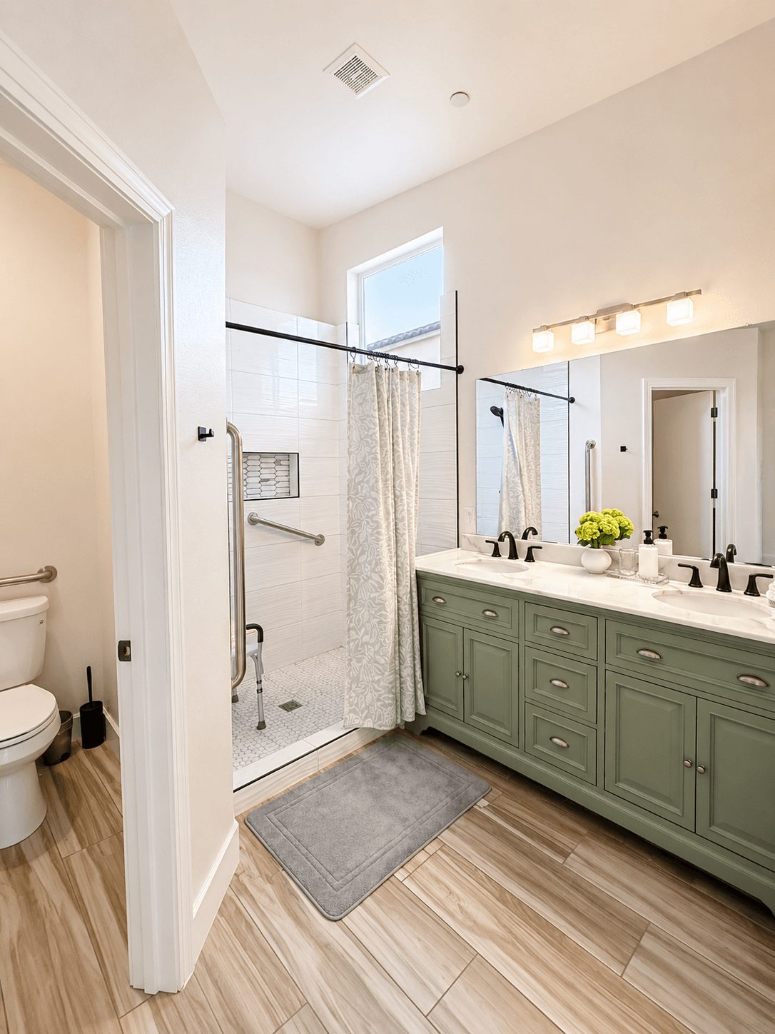 Bathroom with shower, double sink vanity, mirror, and window, light-colored walls, wooden floor, and a gray rug.