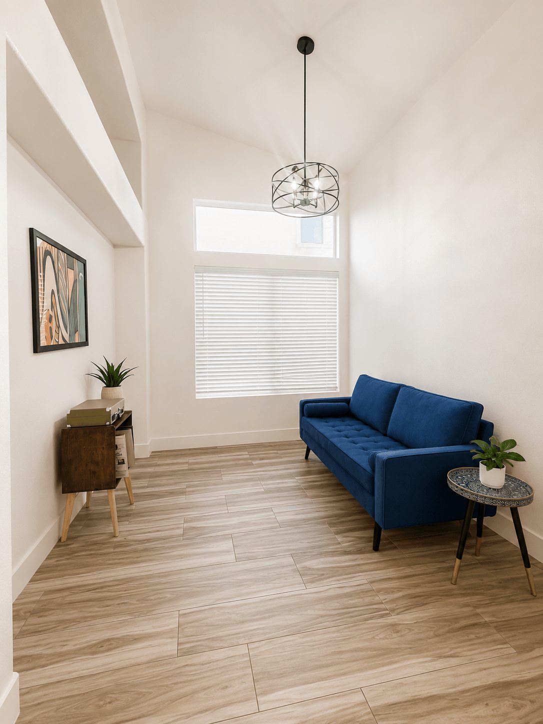 Room with a blue sofa, small side table with plant, window with blinds, and a hanging light fixture.
