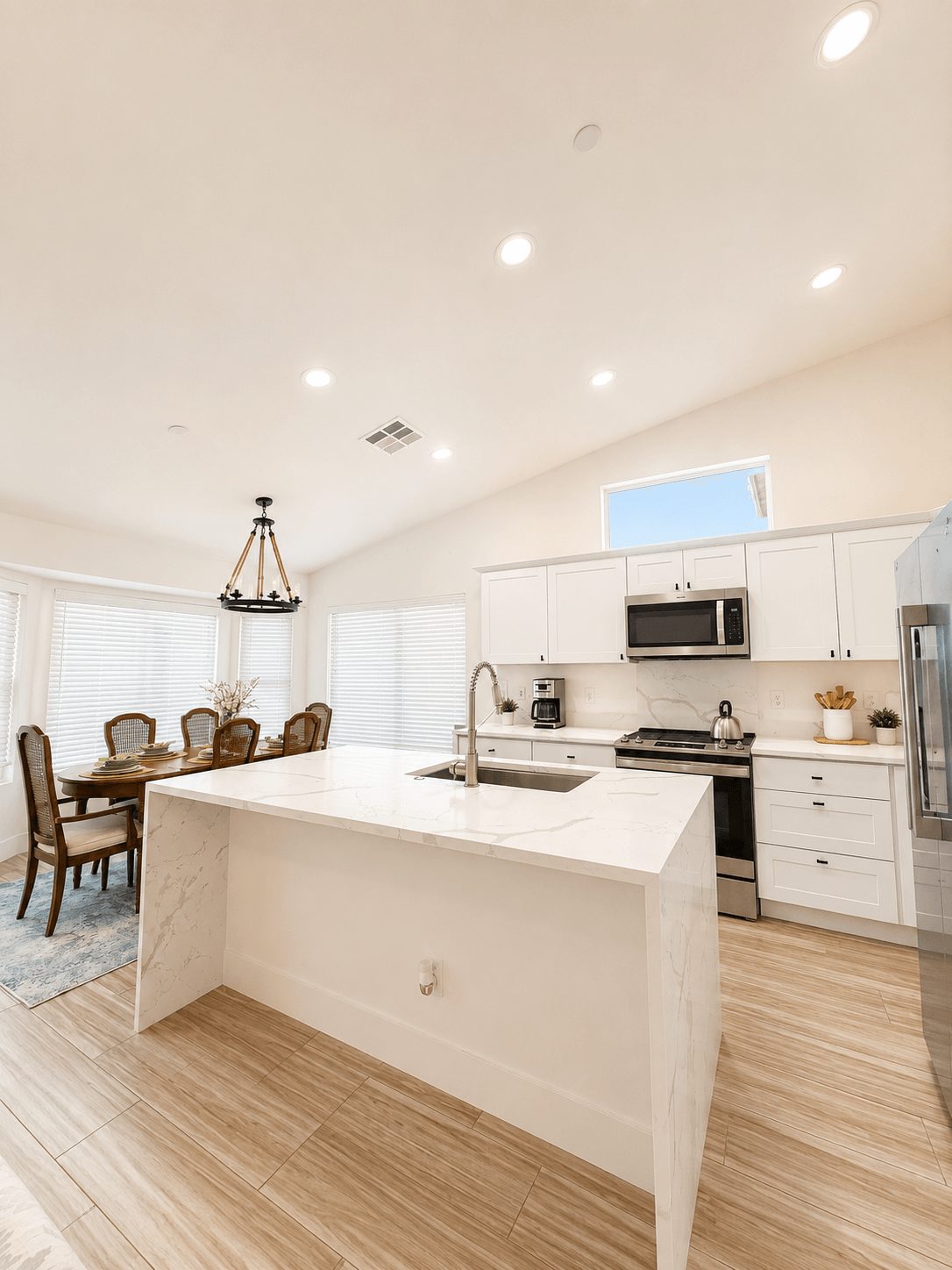 Bright kitchen with white cabinets, island, stainless steel appliances, and a dining area with a chandelier, hardwood floors, and a high ceiling.