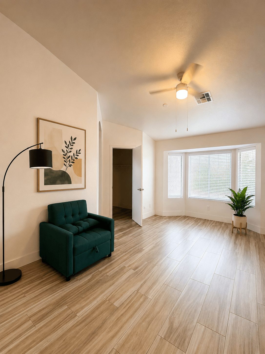 Living room with a green armchair, a floor lamp, a framed artwork, a window with blinds, and a potted plant, hardwood floor.