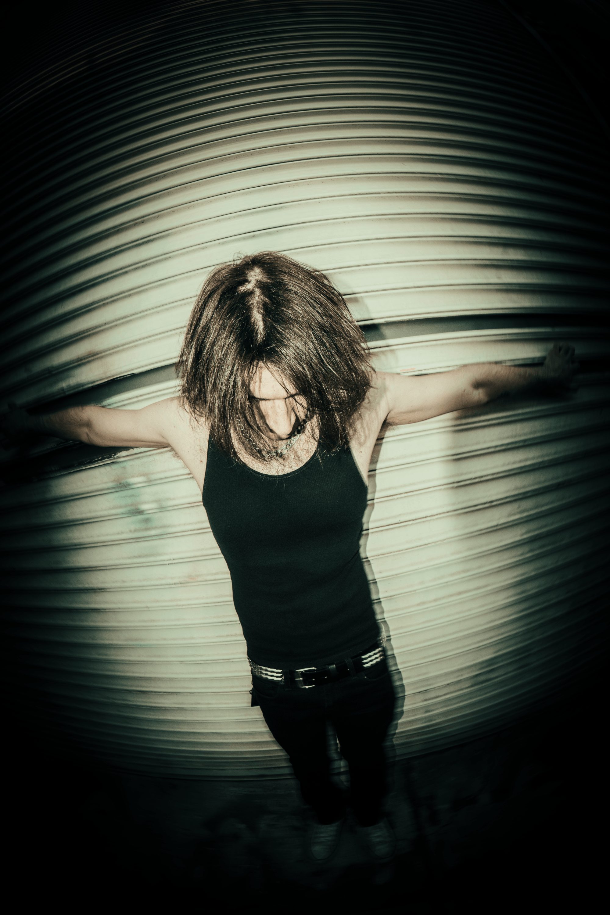 Person with arms outstretched, head down, standing in front of a curved, ribbed metallic surface, wearing a black top.