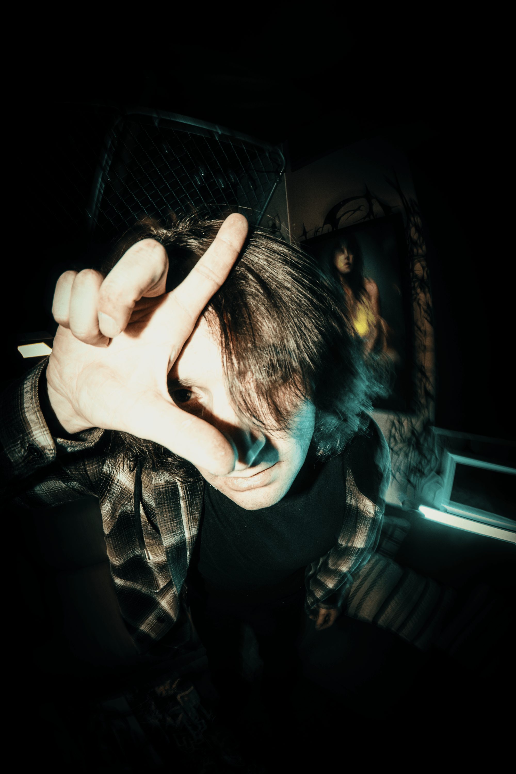 Person with dark hair and blue streaks making a hand gesture in a dark room with metal structures behind.