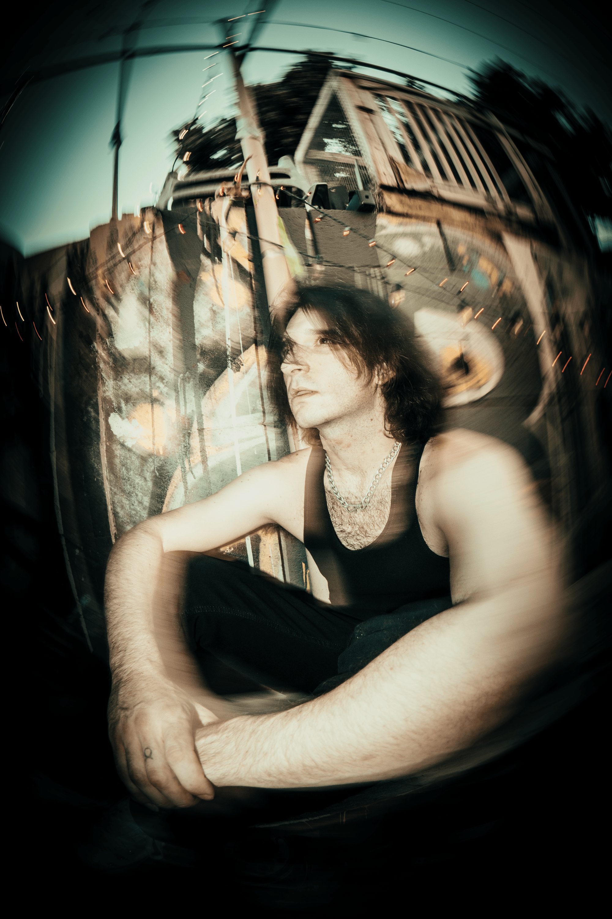 Woman with dark hair and sleeveless top sitting outdoors, with a blurred background of structures and sky.