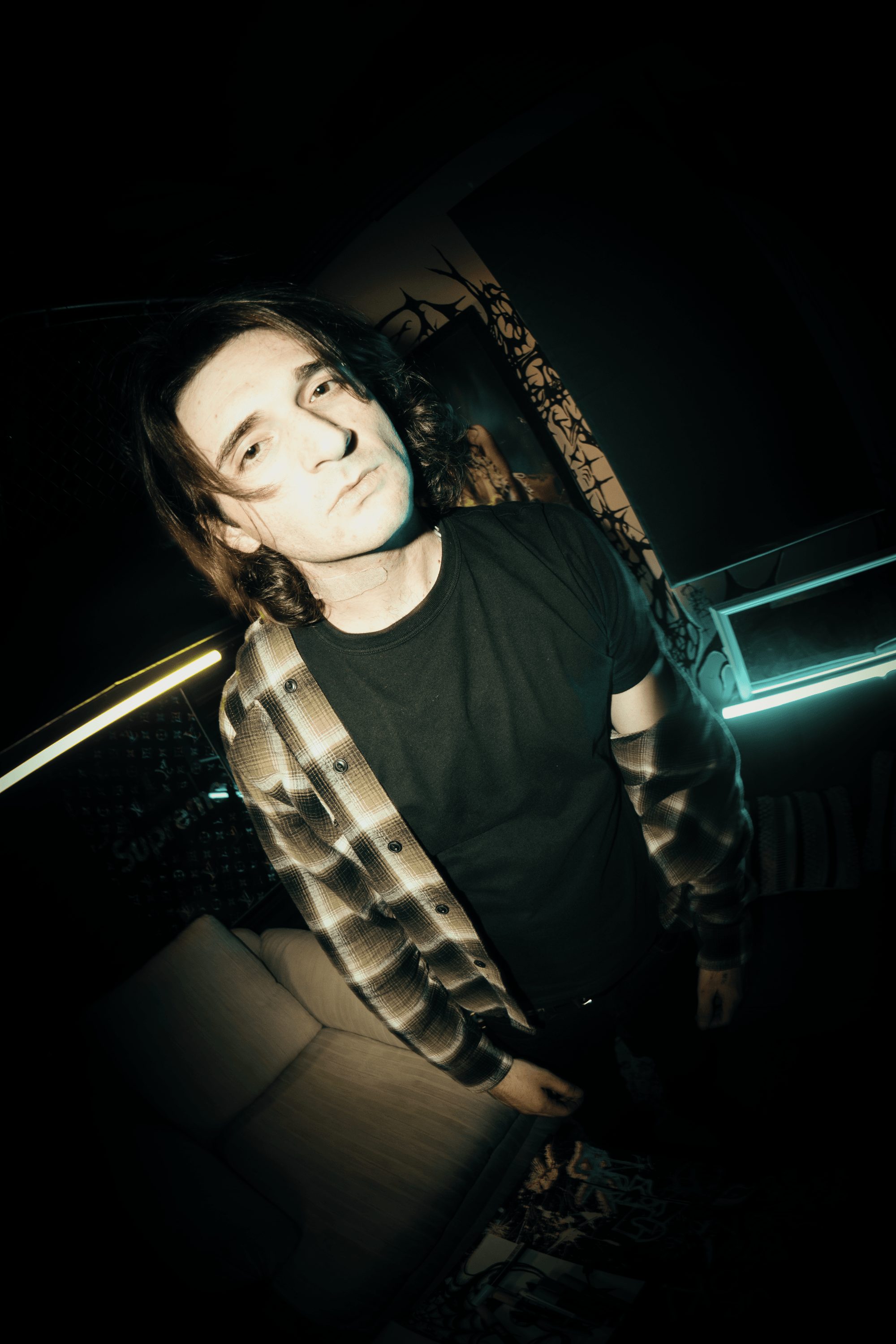 Young person with dark hair and light skin, wearing a black shirt and checkered jacket, in a dimly lit room.