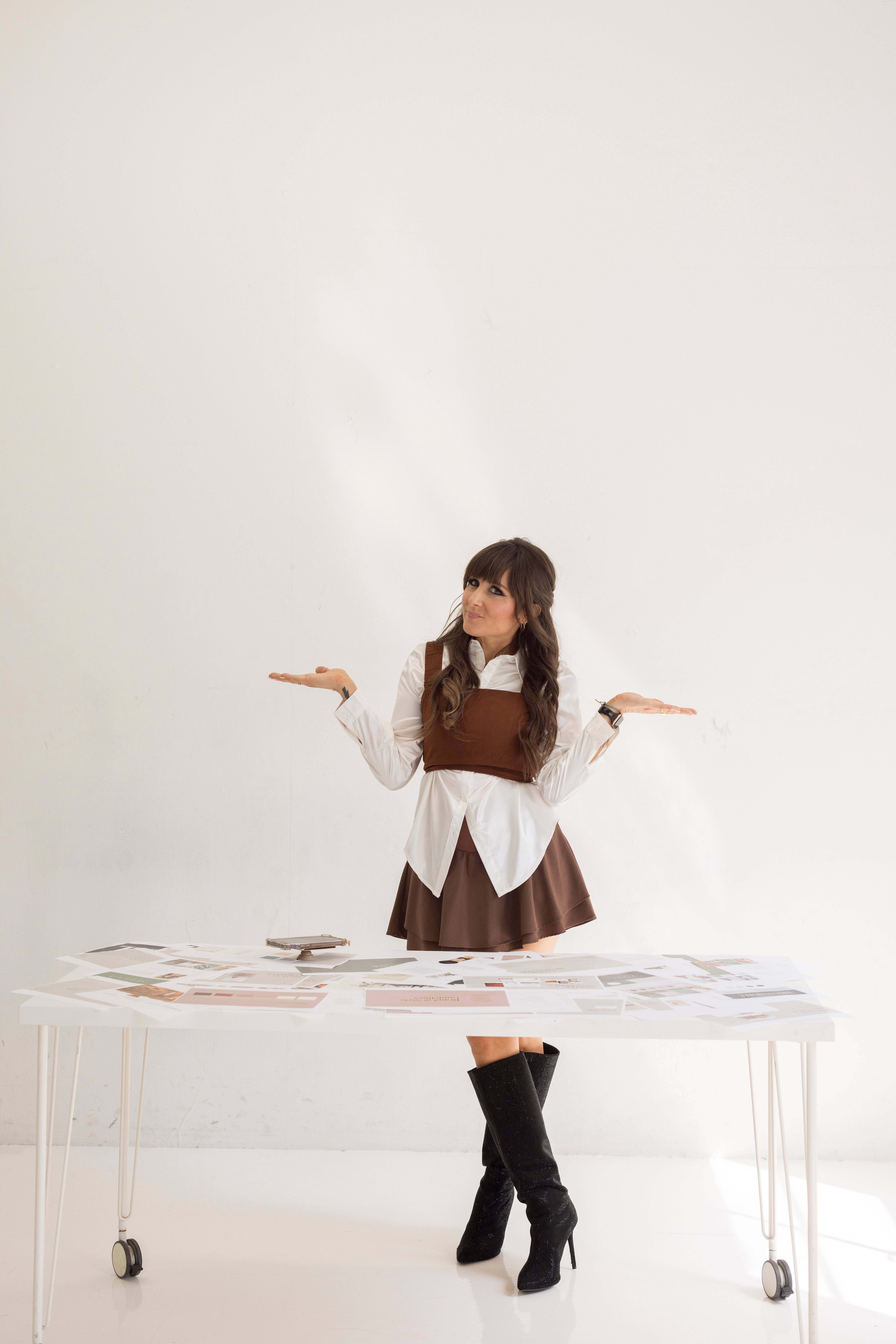 Young woman with long hair in a brown and white outfit stands behind a table with papers, arms raised, in a bright room.