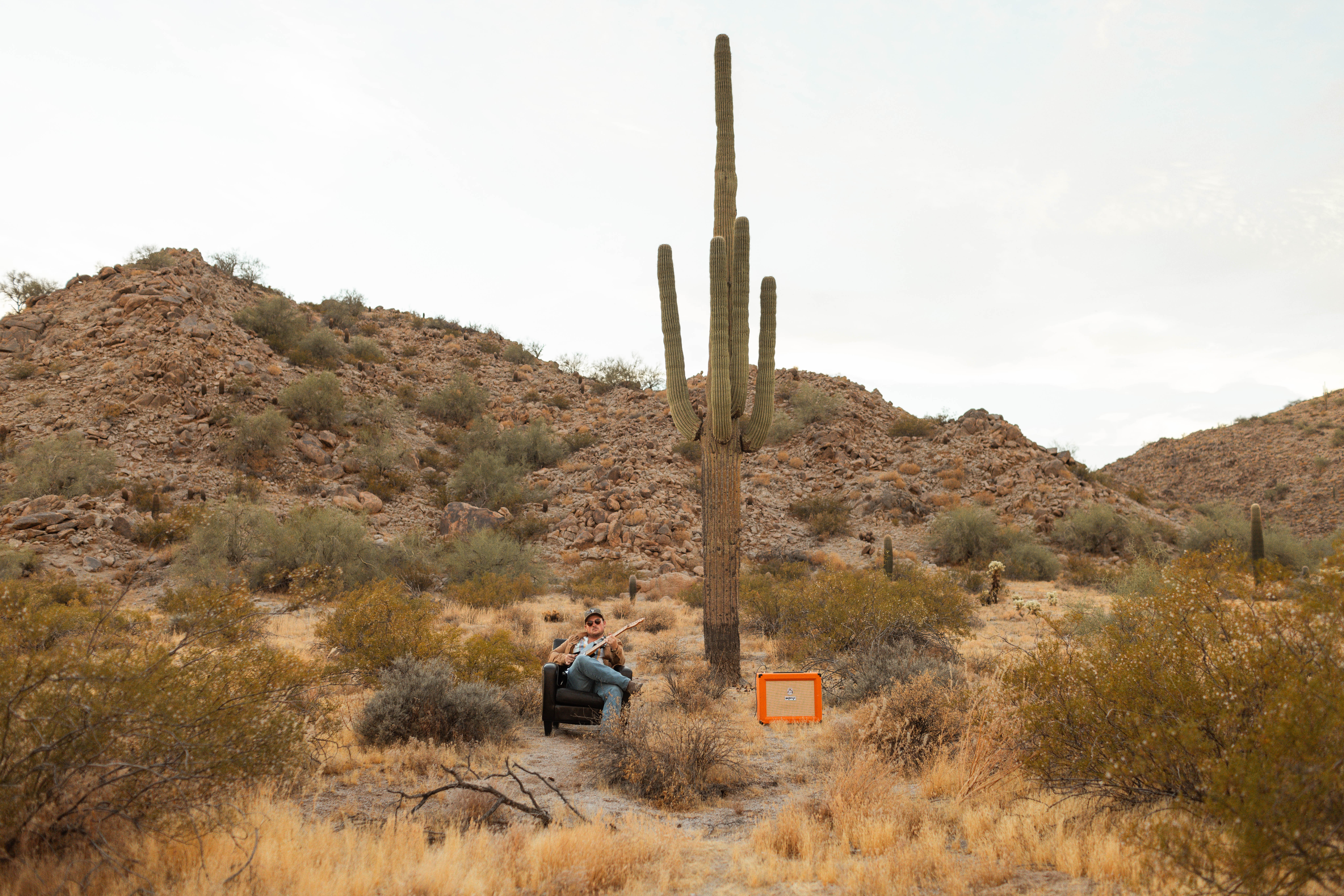 Person with backpack standing near a tall saguaro cactus in a desert landscape with hills and sparse vegetation