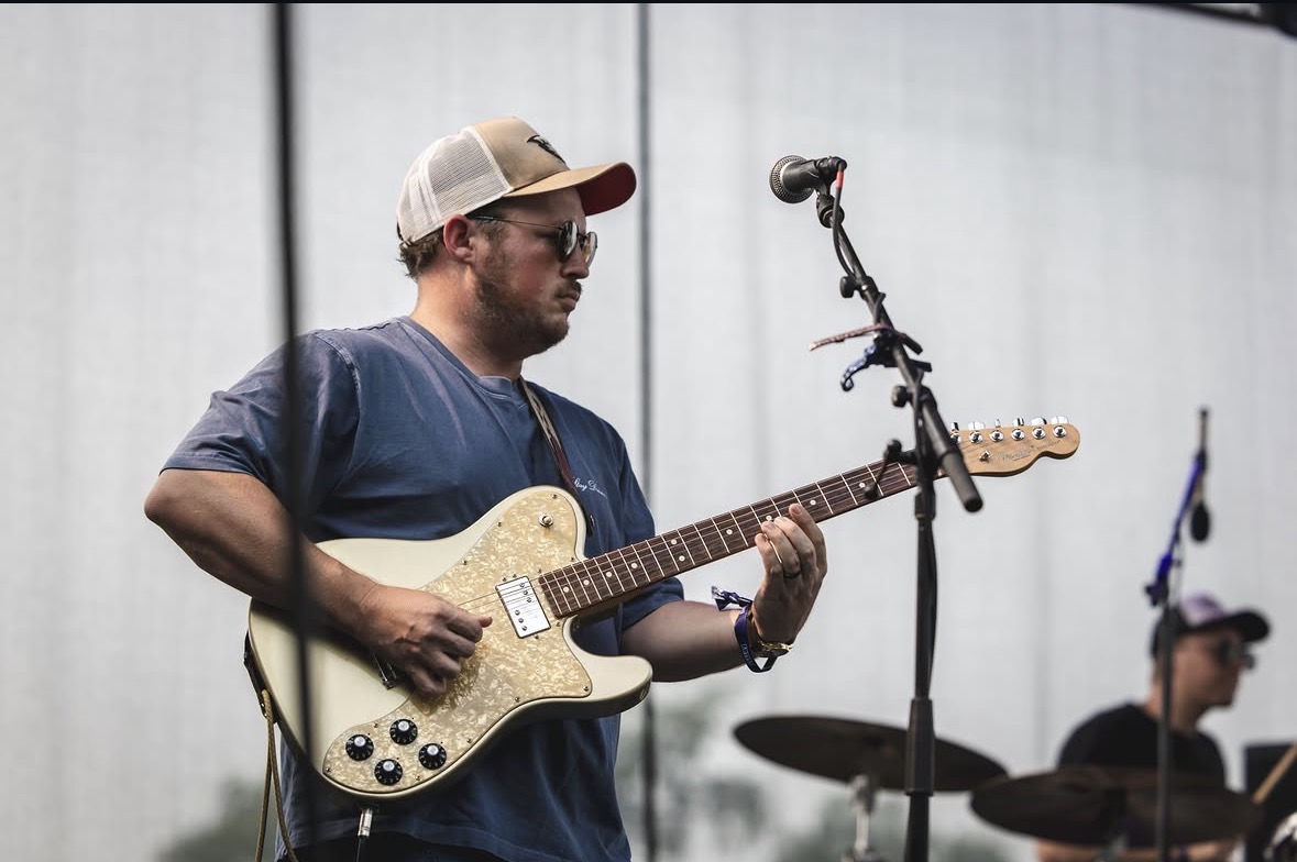 Person playing electric guitar on stage with microphone stand nearby, background shows a plain wall, another musician in the background.