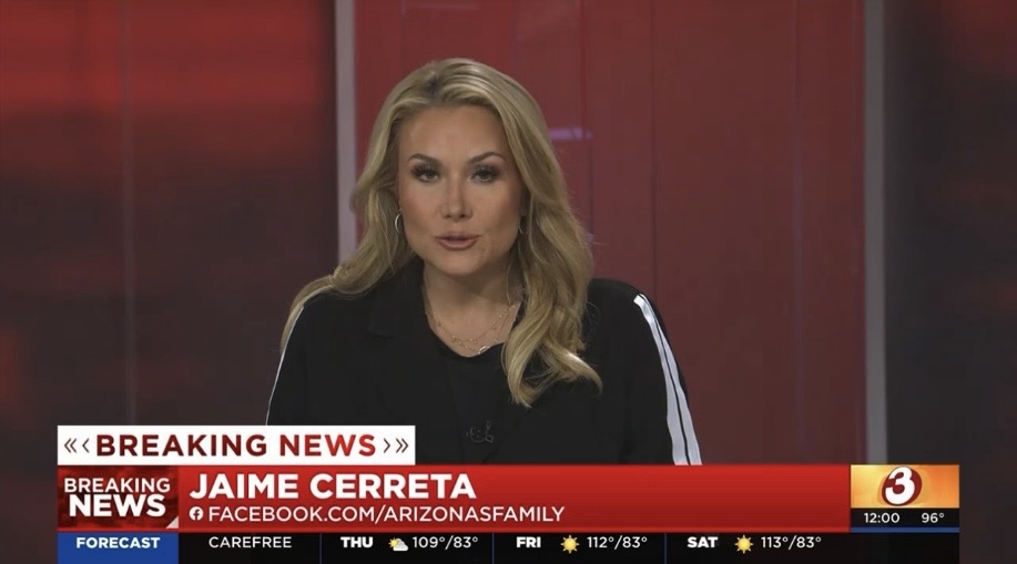 Female news anchor with blonde hair wearing a black top, sitting at a news desk with a red background.