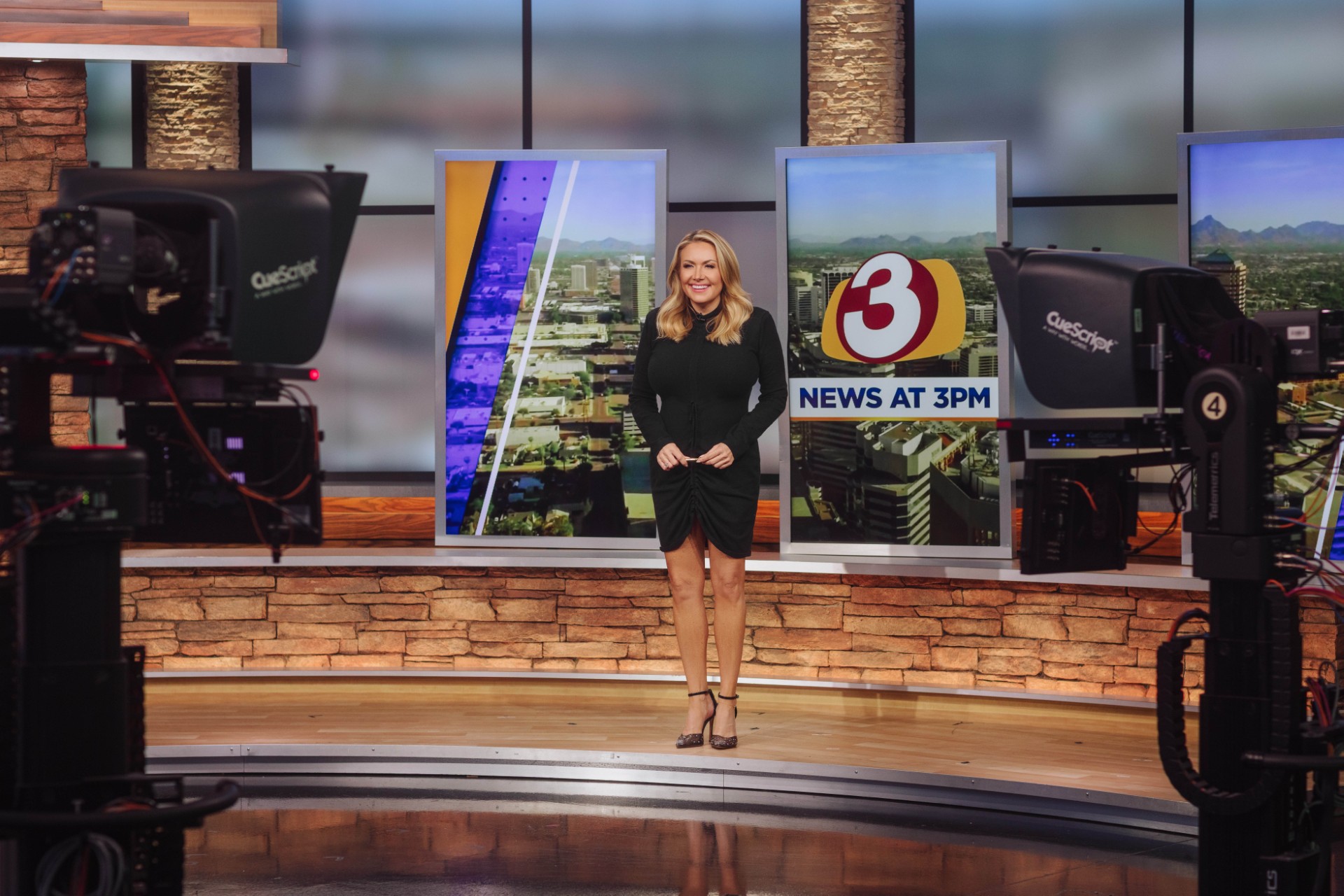News anchor standing on a studio set with screens and cameras, smiling, with a brick wall background.