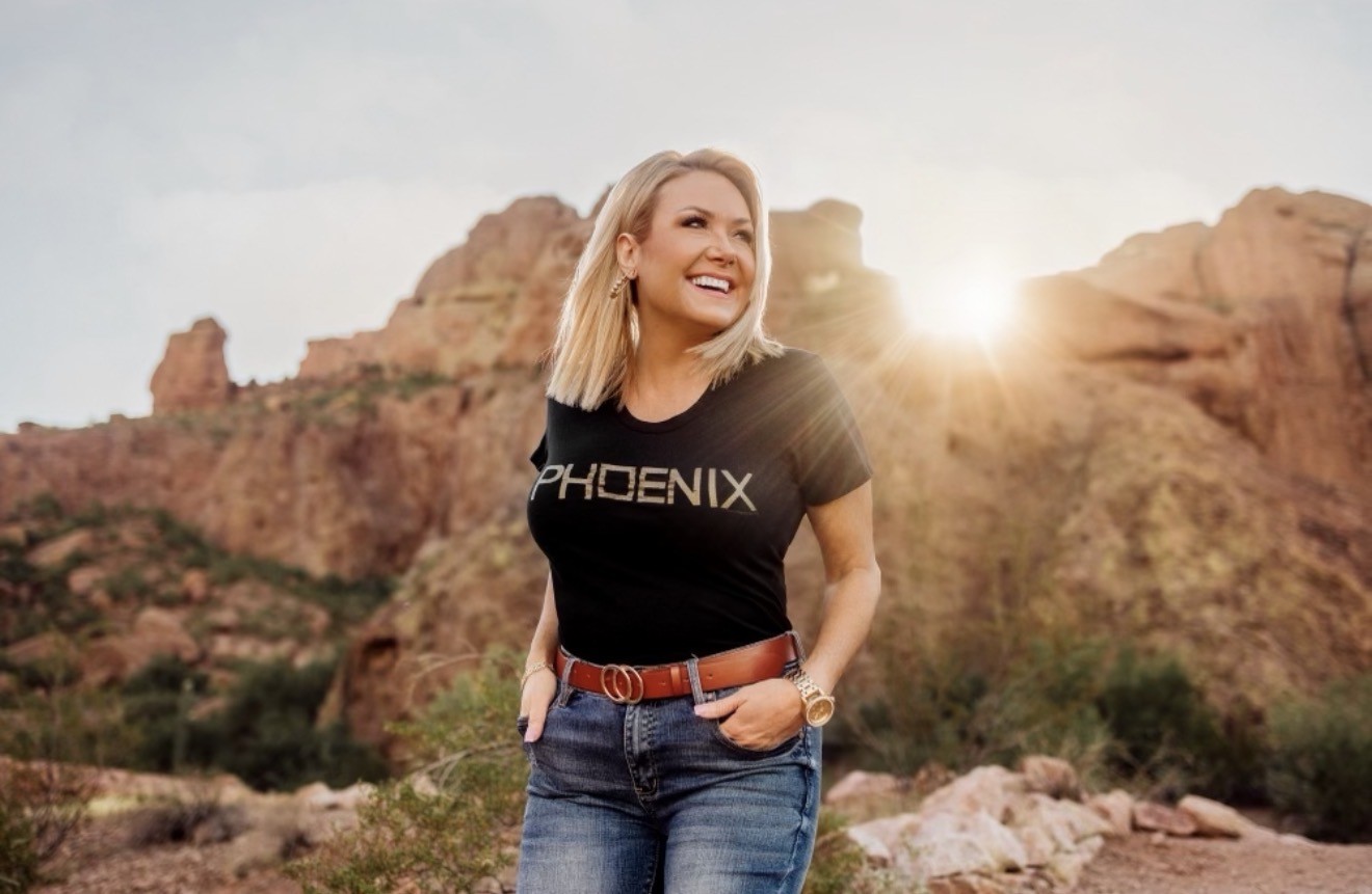 Woman smiling outdoors with rocky mountains in background, sunlight behind her, wearing a black t-shirt and jeans.