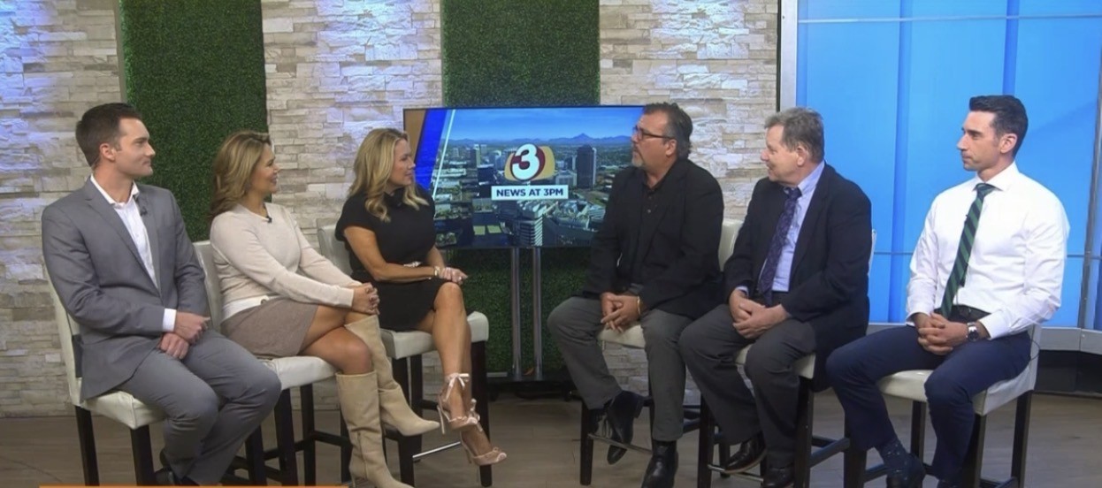 Six people seated in a studio, engaged in conversation, with a large screen behind them displaying graphics.