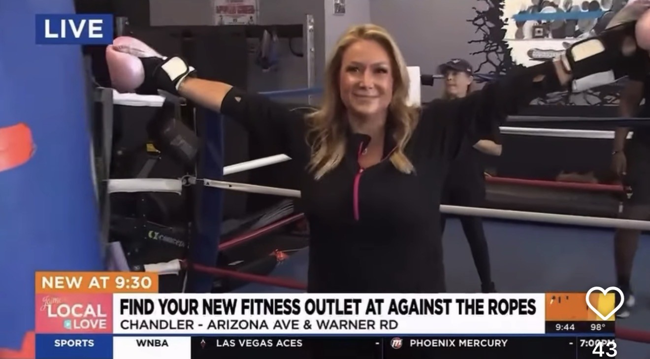 Woman in a gym with boxing gloves, smiling with arms outstretched, boxing ring in background, fitness activity setting.