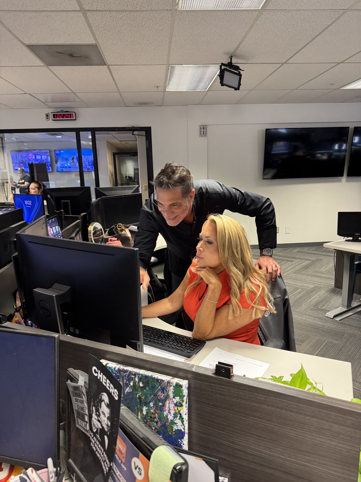 Man leaning over a seated woman at a desk with multiple computer monitors in an office, both looking at the screens.