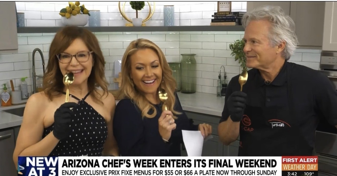 Three people in a kitchen holding forks with food, smiling, with a white tiled wall and plants in the background.