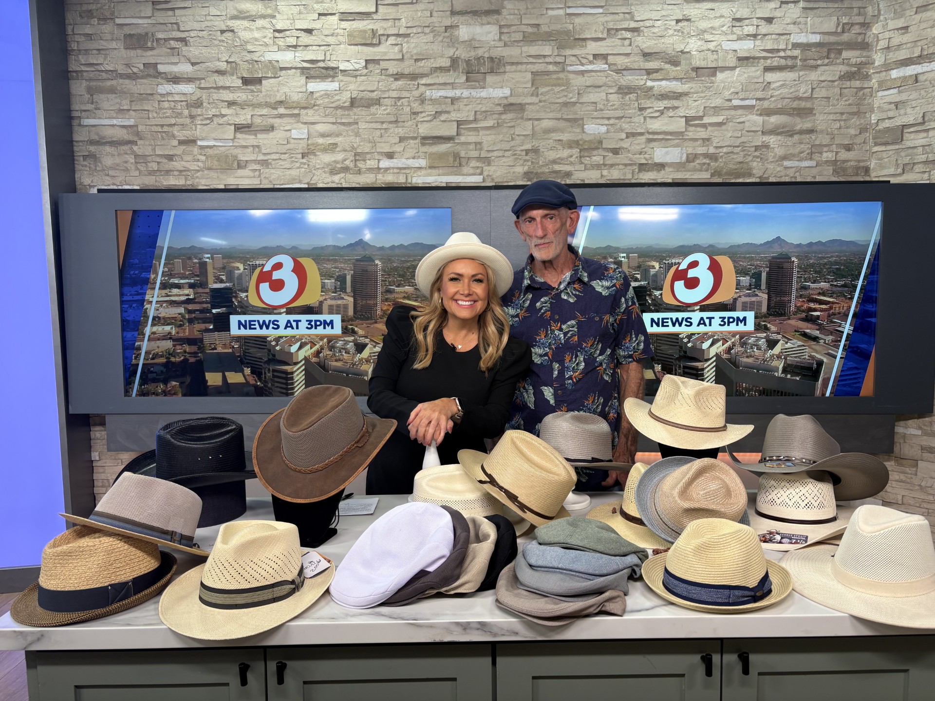 Two people stand behind a table with various hats, in a news studio with cityscape background, smiling.