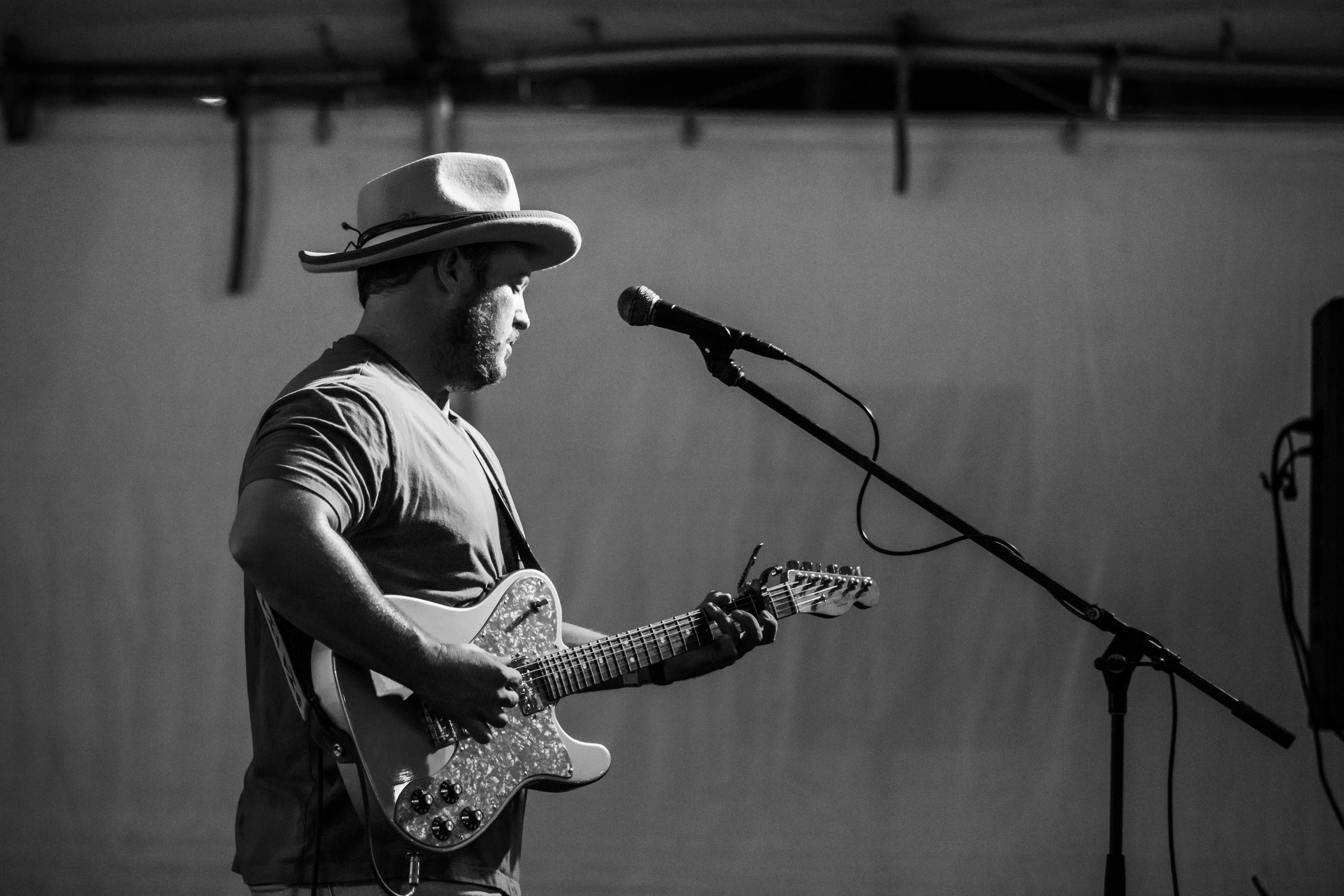 Man wearing a hat playing an electric guitar on stage with a microphone, in black and white.