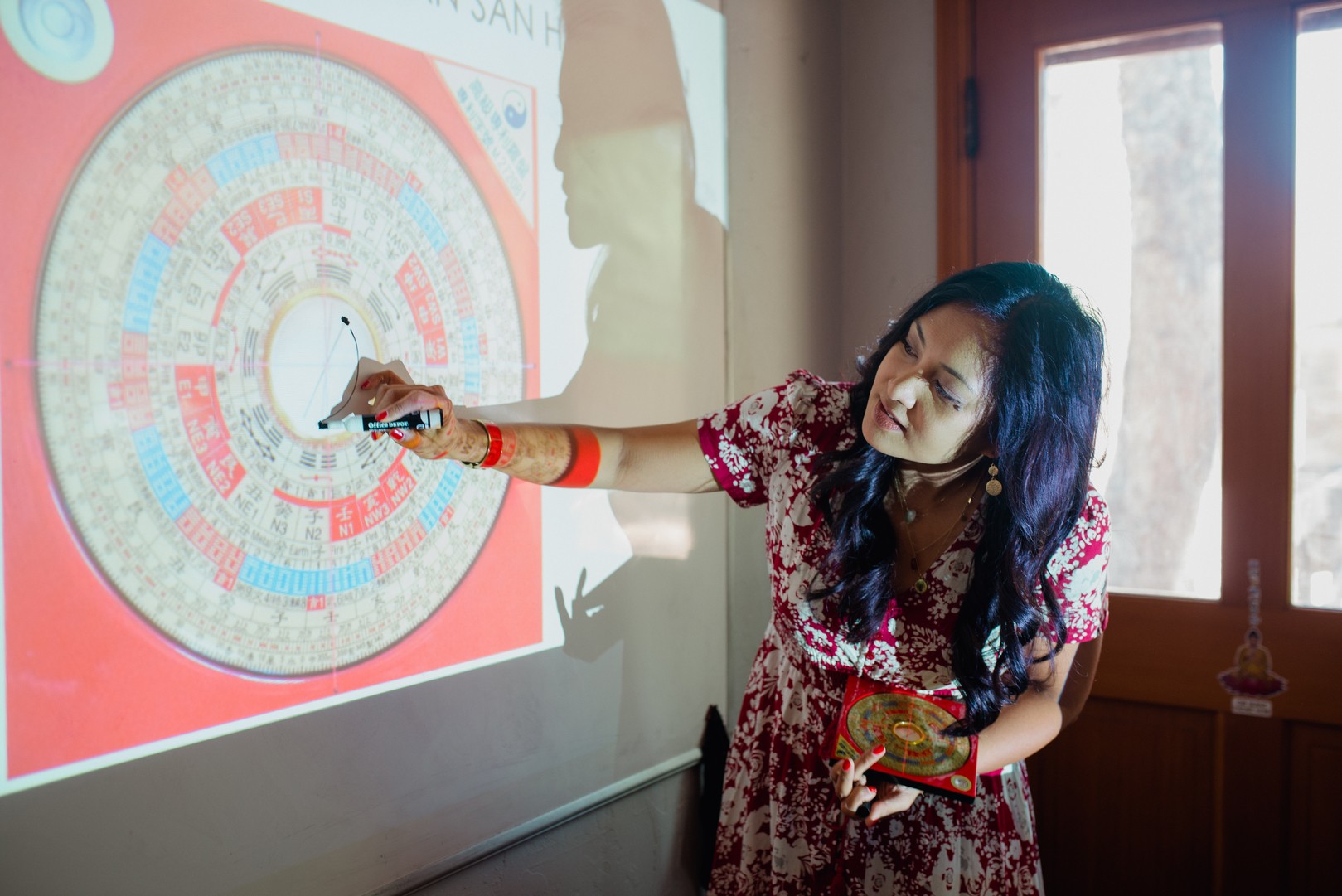 Woman pointing at a colorful circular diagram projected on a wall, holding a pointer and a small object, in a room with a window.