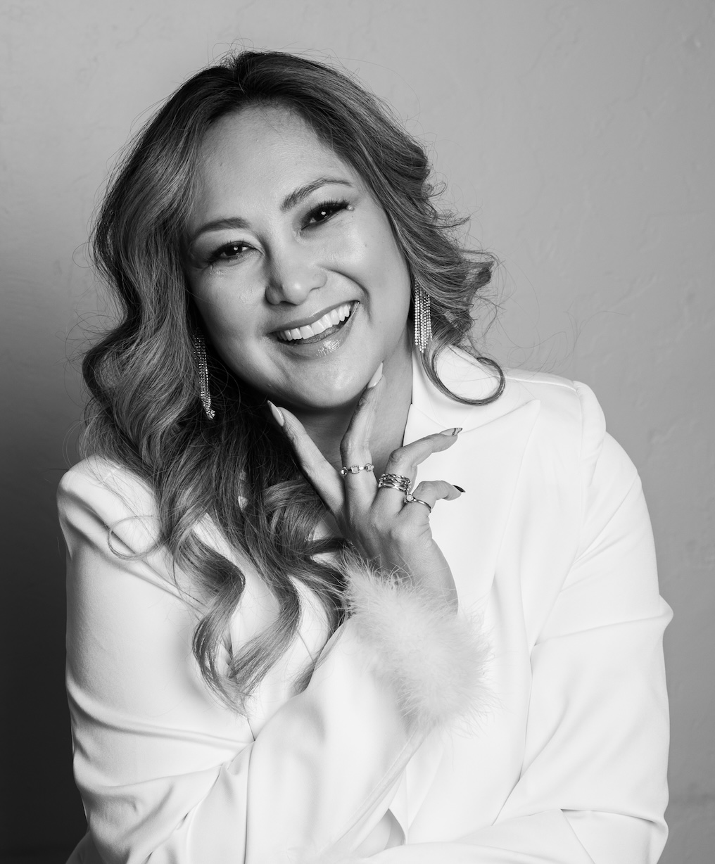 Woman with wavy hair smiling, wearing a white blazer, touching her face with her hand, in black and white.