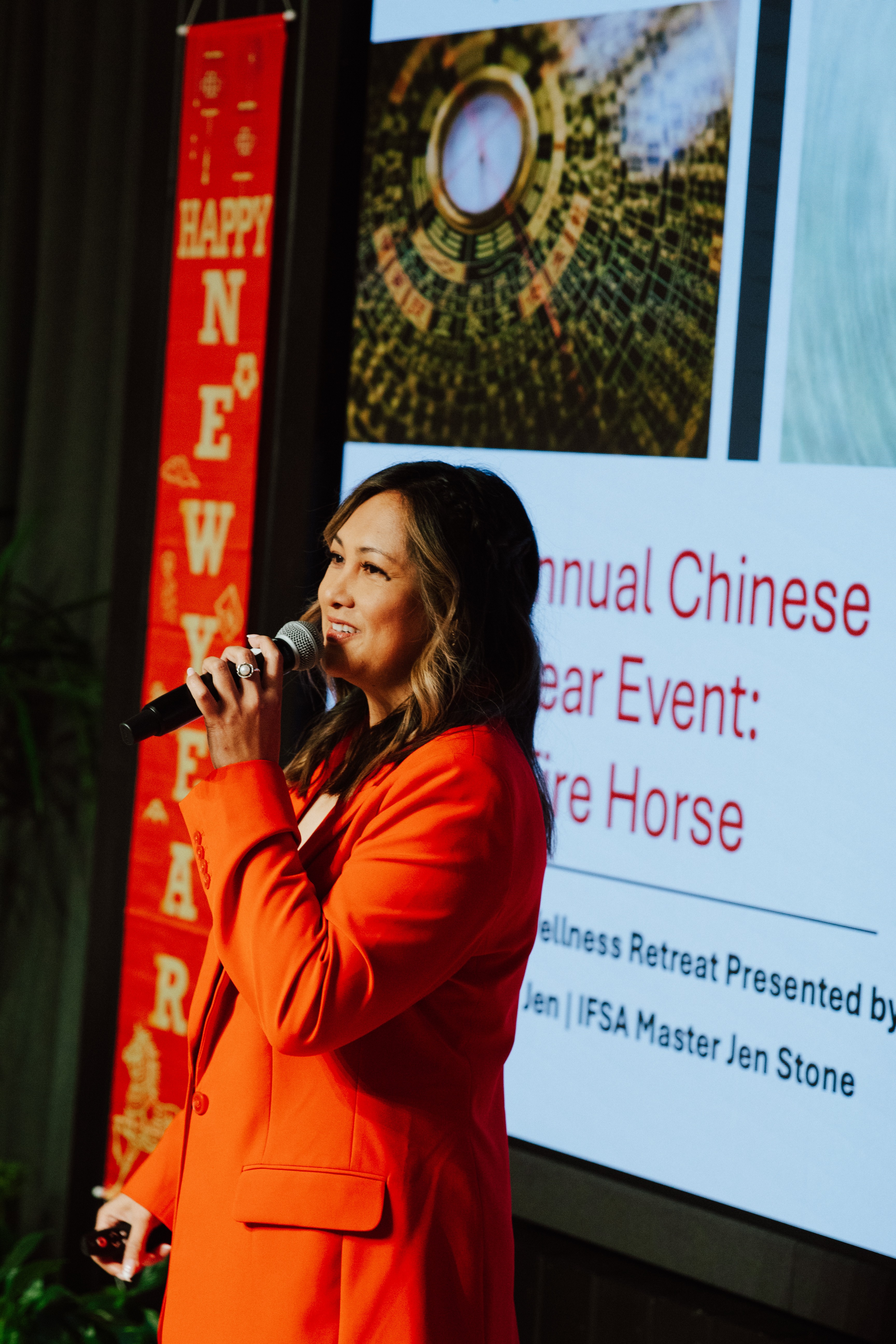 Woman in red blazer speaking into microphone at event with screen and red banner in background.
