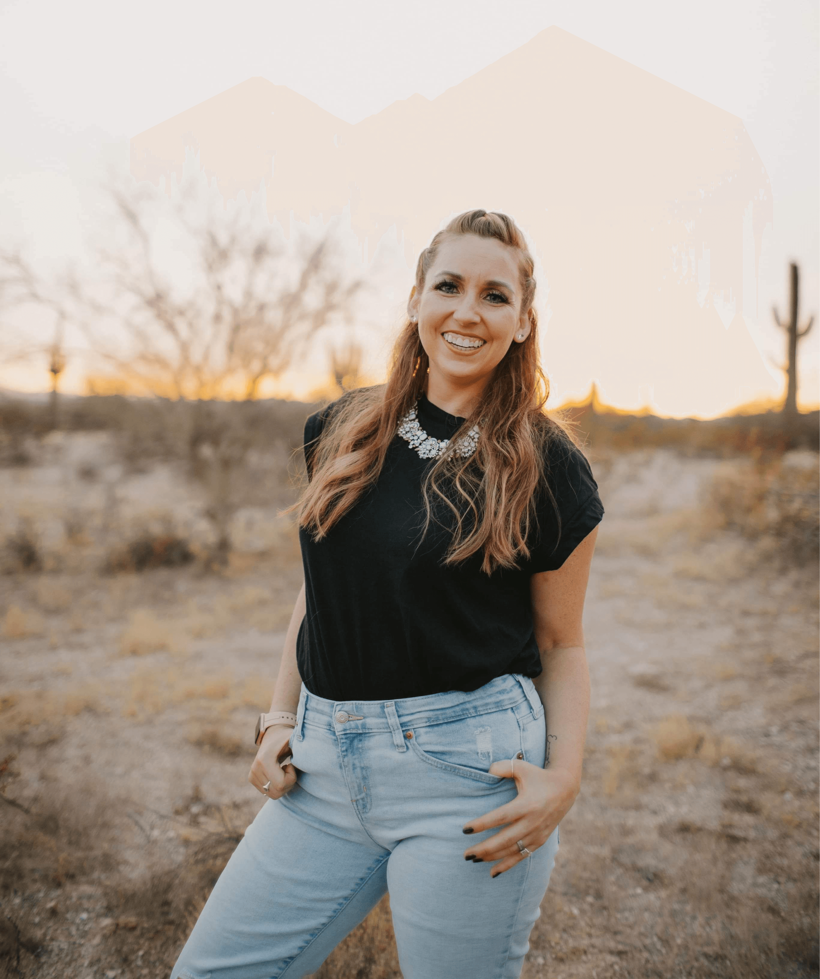Smiling woman in black top and light jeans standing outdoors in desert landscape with cacti and trees, sunset in background.