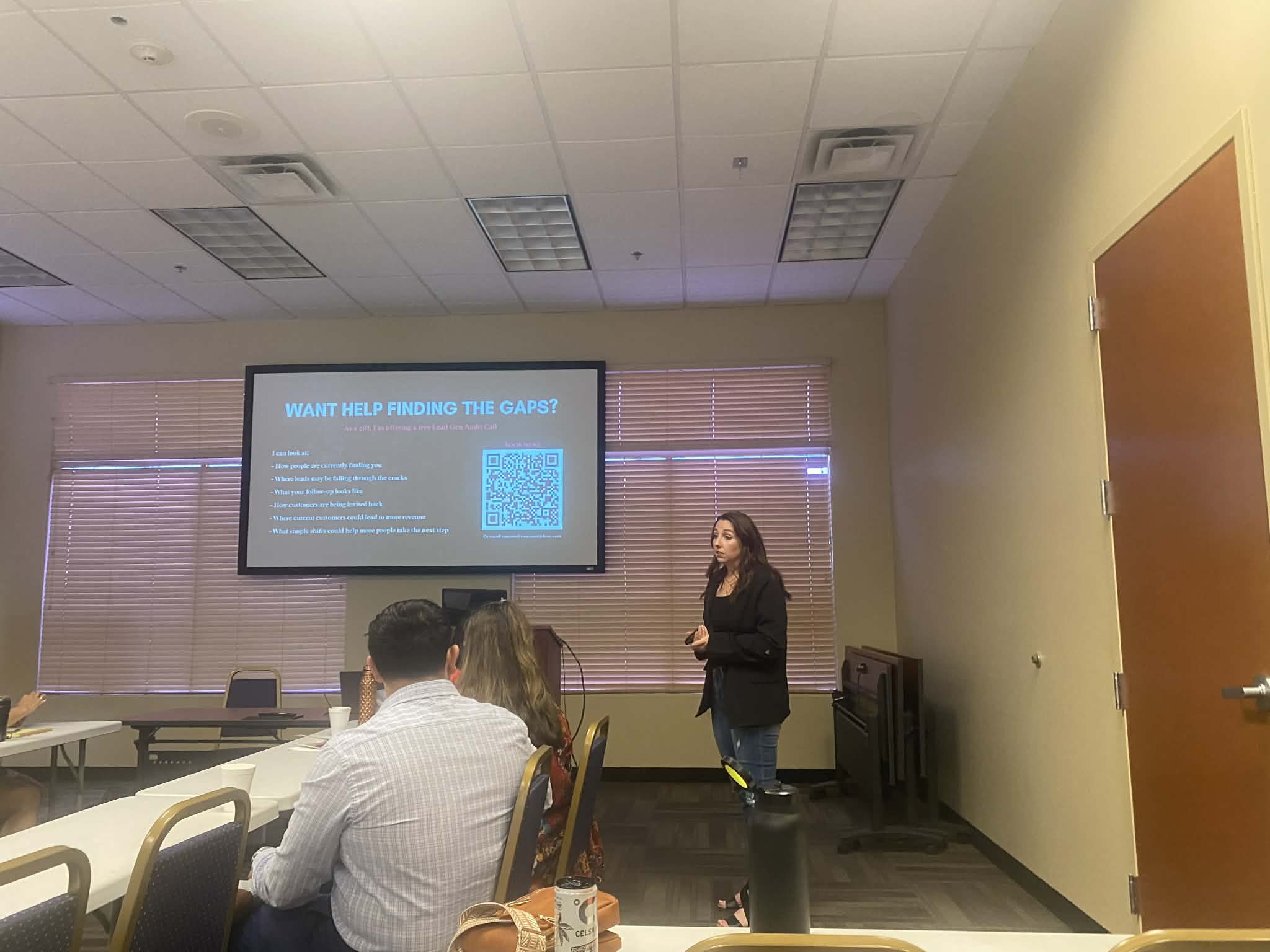 A woman presents in a classroom with a projected slide, seated audience, and windows with blinds behind her.
