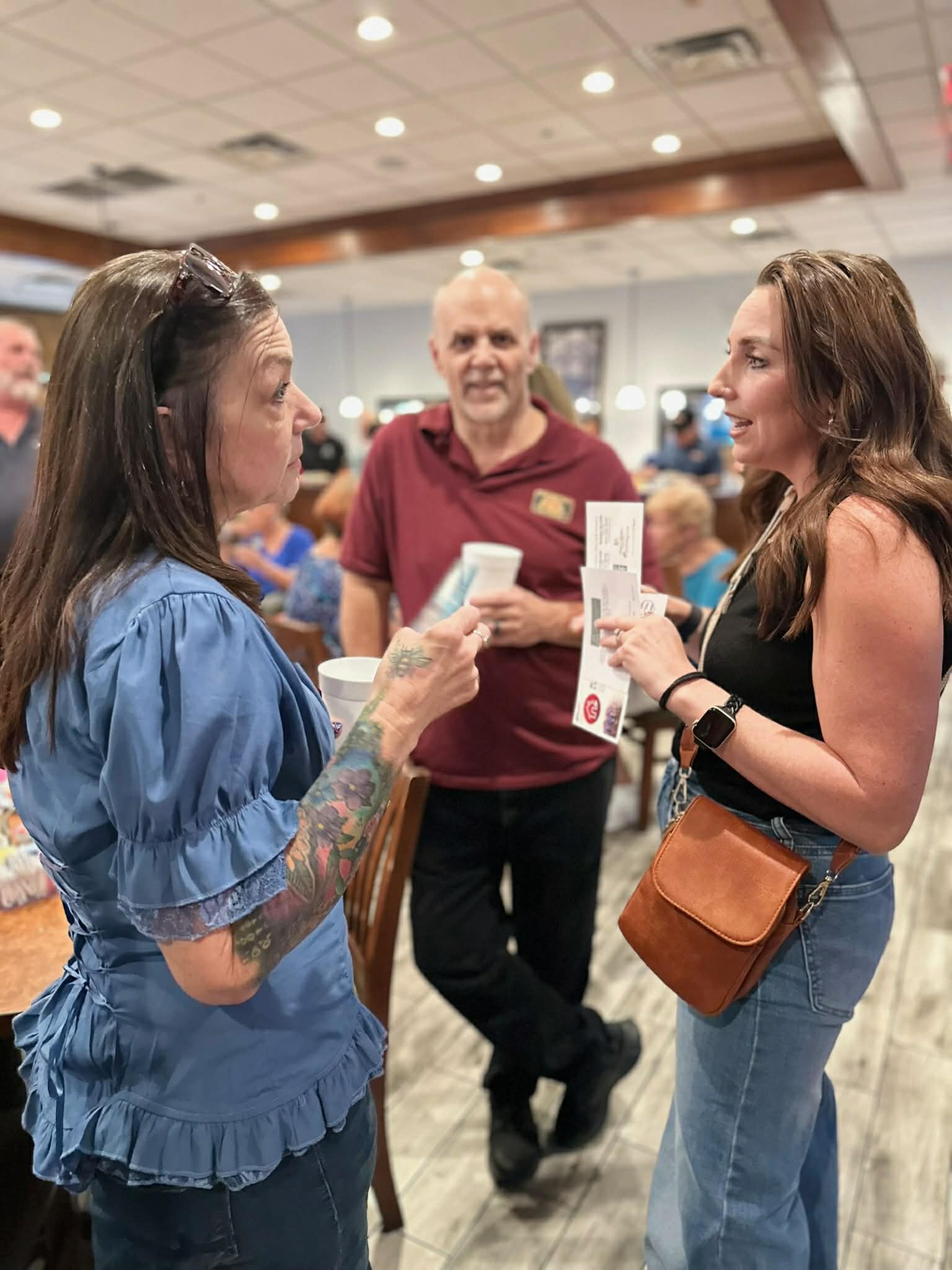 Two women and a man engaged in conversation indoors, with people and tables in the background.